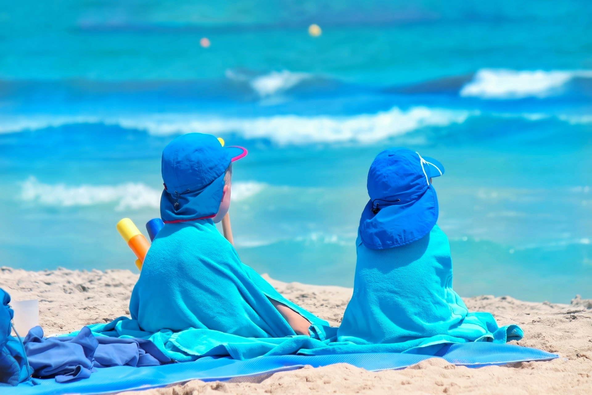 青いビーチタオルに座る二人の子供たちが青い海を見つめている