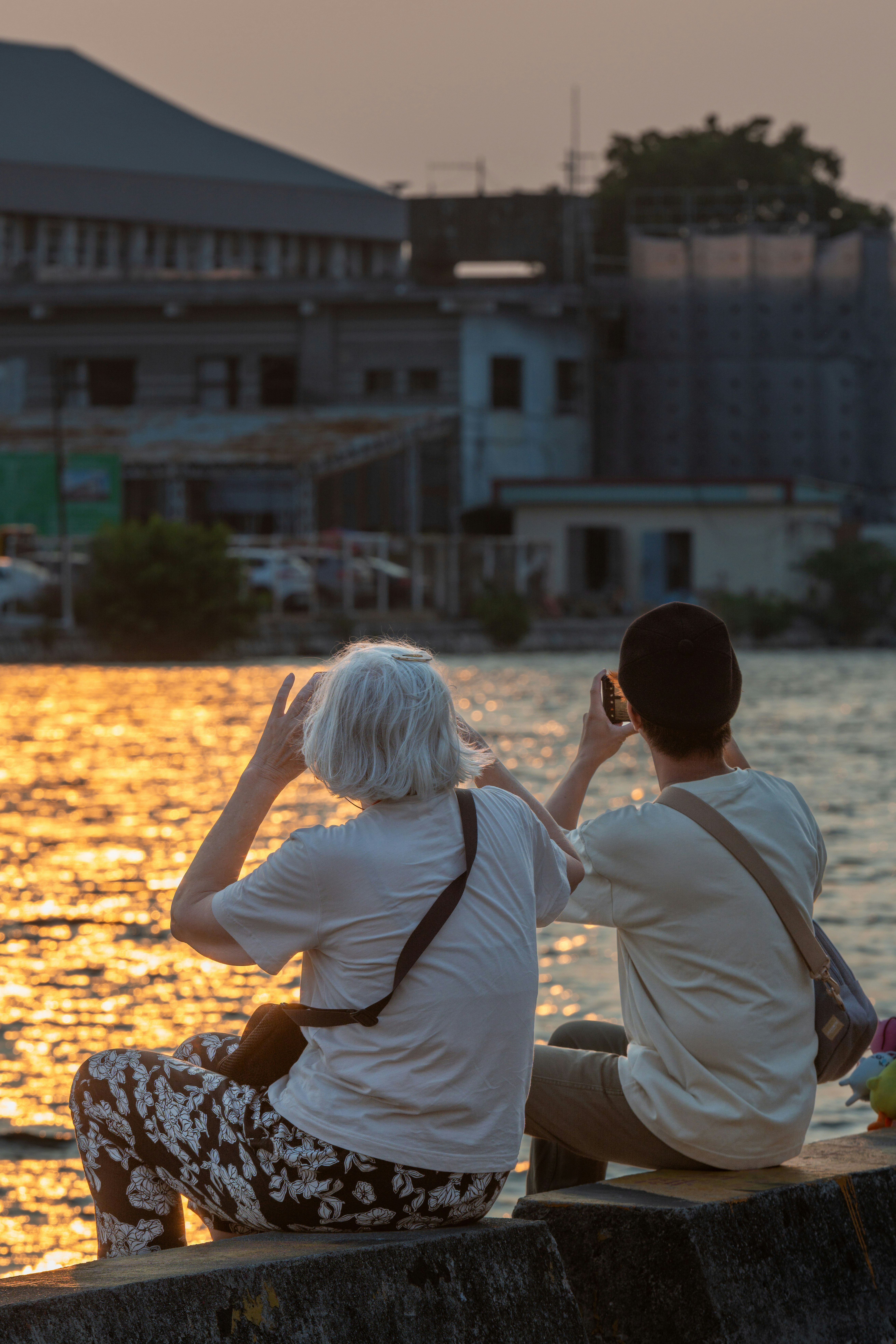 夕日を背景に川のほとりに座る二人の人々 白髪の女性と若い男性がスマートフォンで写真を撮っている