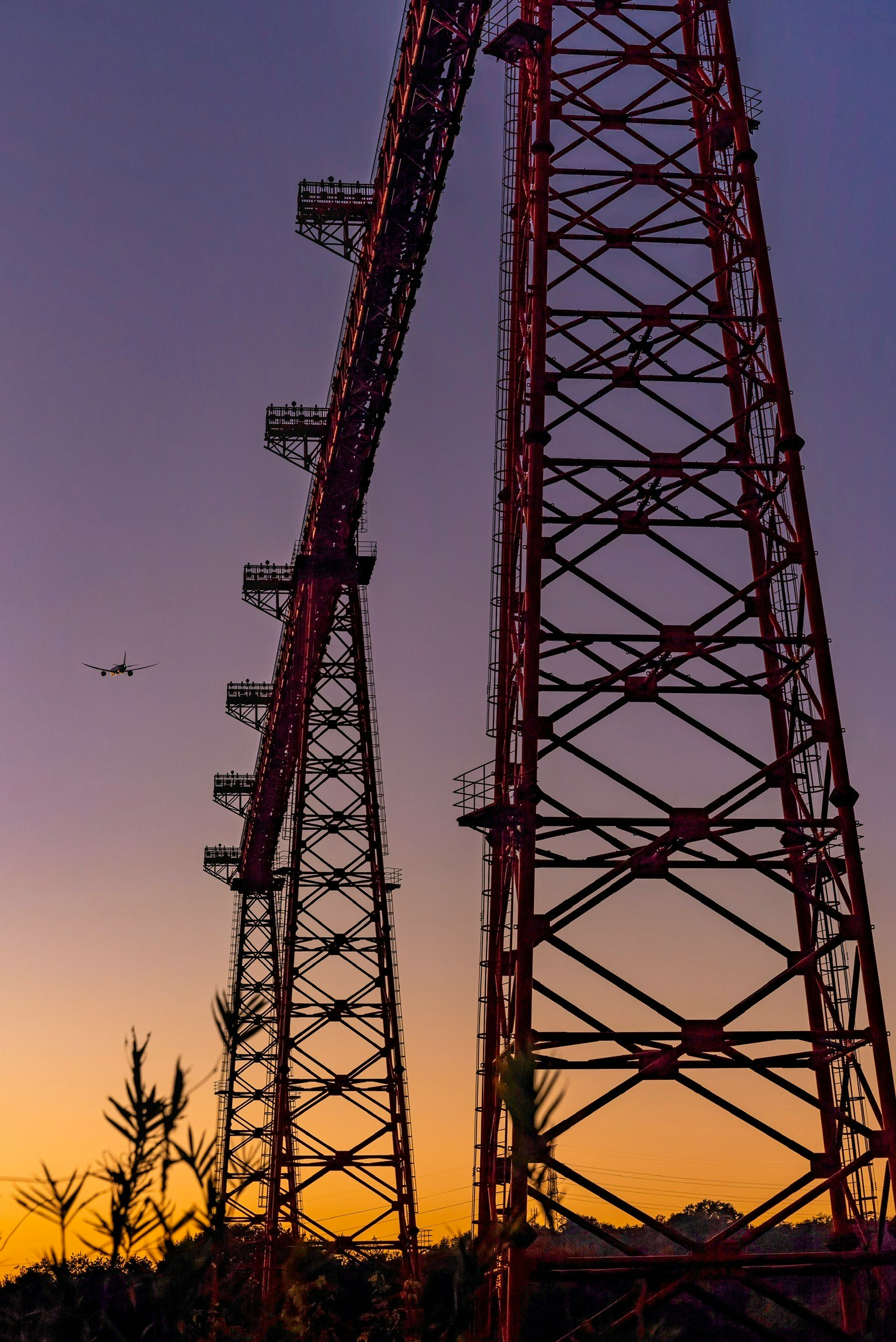 Zwei Stahlmasten silhouettiert gegen einen Sonnenuntergang mit einem Flugzeug