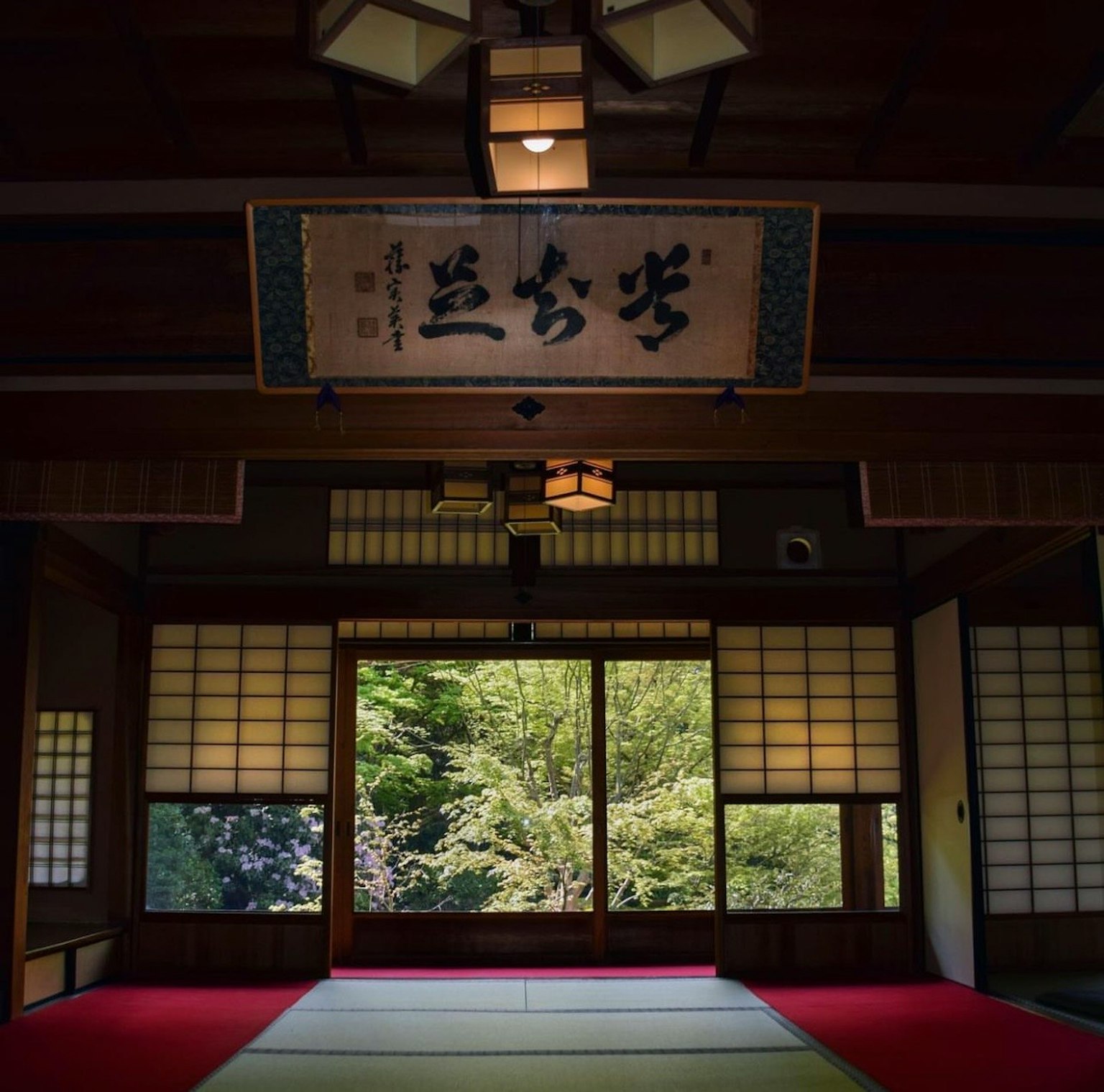 传统日本房间的内部，采用障子窗户，展现绿色景色，地面铺有红色榻榻米，上方悬挂着书法卷轴
