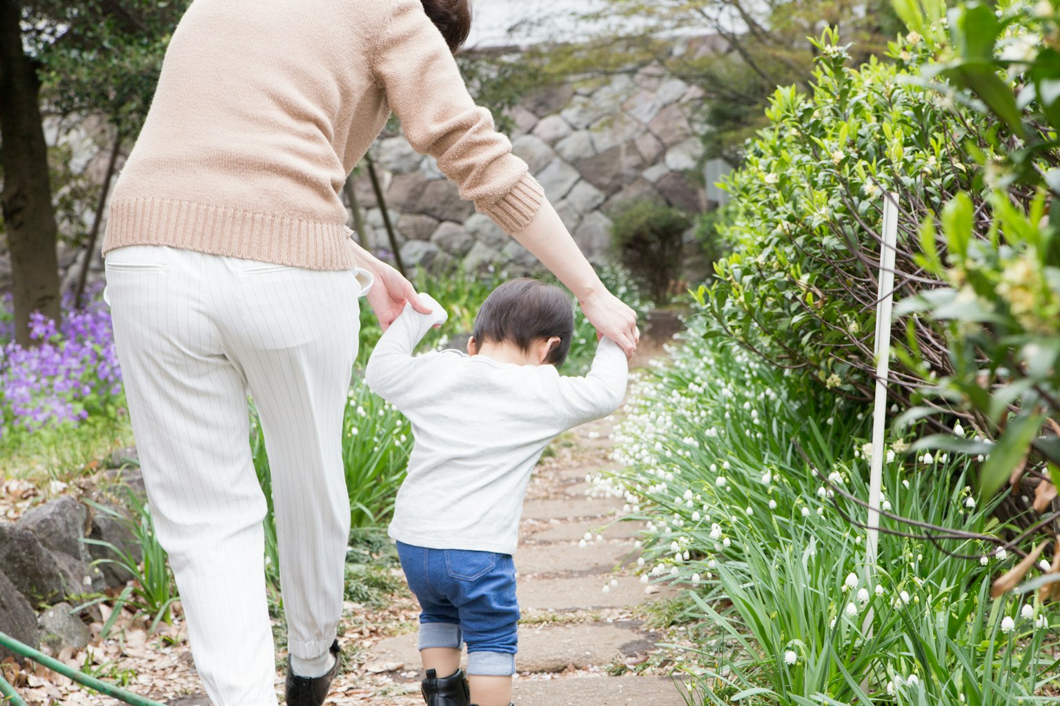 Una madre che tiene per mano il suo bambino mentre camminano lungo un sentiero verde circondato da fiori