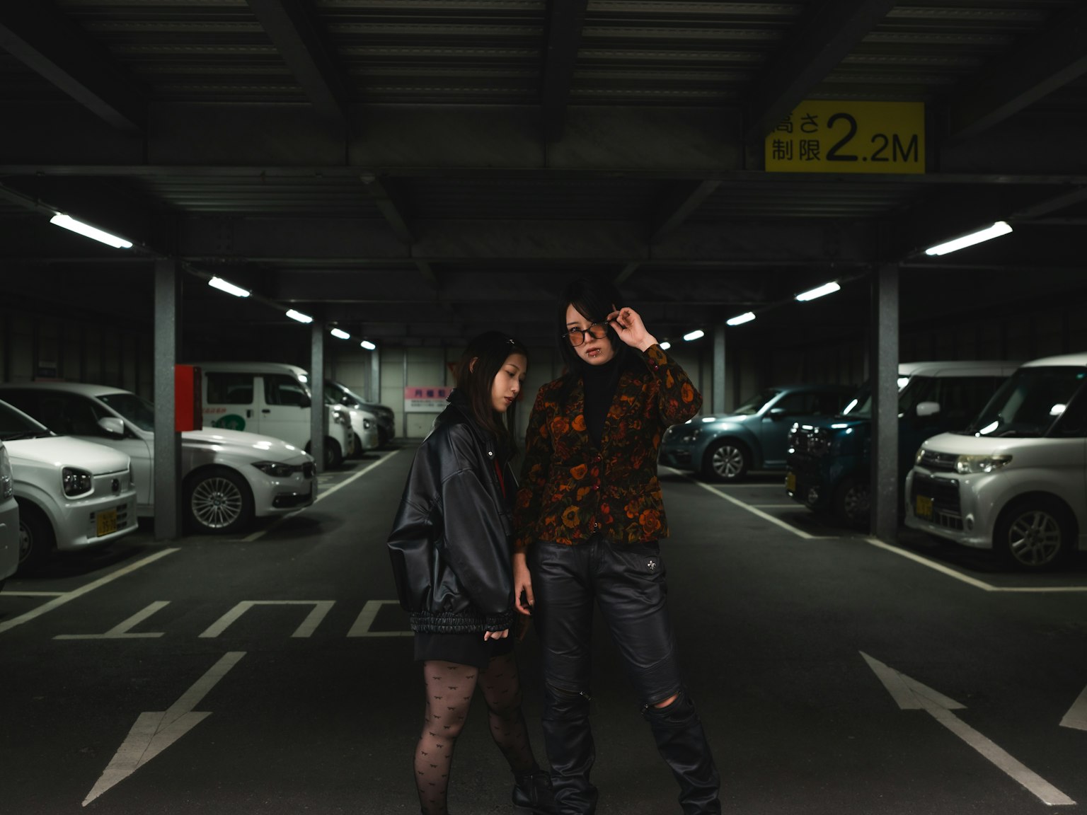 在停车场摆姿势的两位女性 一位穿着黑色夹克 另一位穿着彩色毛衣 周围停着白色汽车