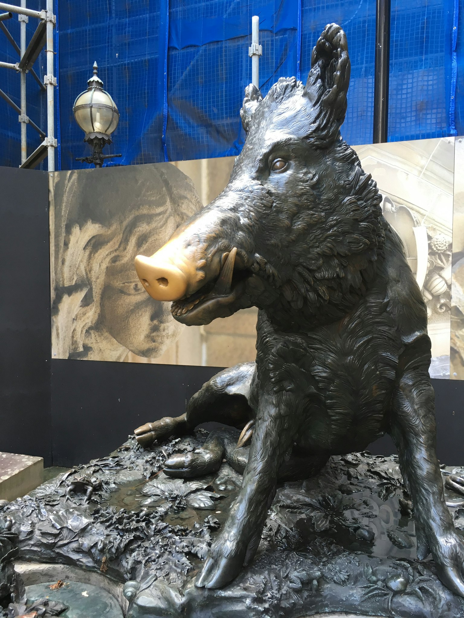 Scultura di cinghiale in bronzo seduta con uno sfondo metallico e un lampione visibile