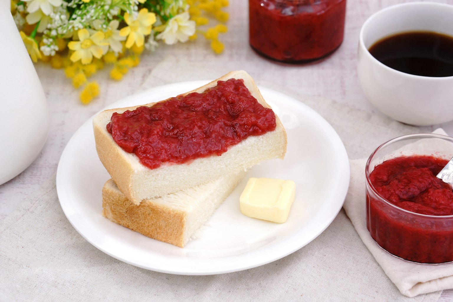 Une assiette avec du pain tranché garni de confiture rouge et d'une noisette de beurre accompagné de café et d'un pot de confiture avec des fleurs décoratives