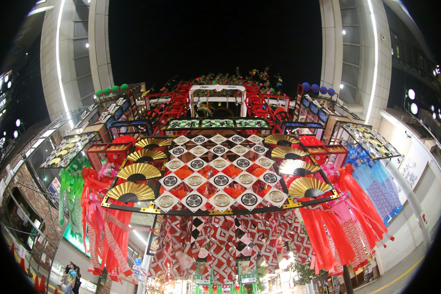View from below a festival stall at night featuring colorful decorations and traditional fans