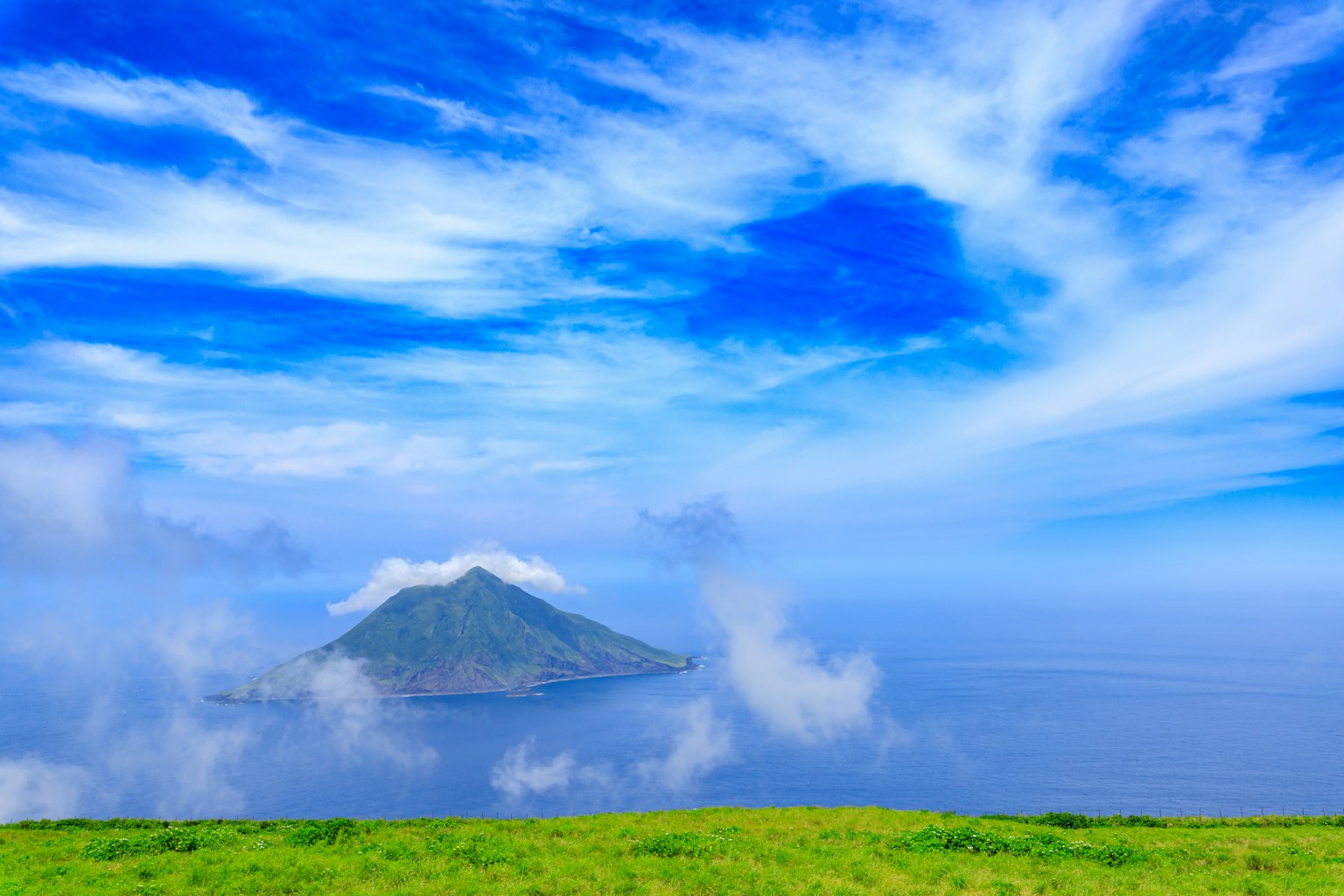 Island surrounded by blue sky and clouds with green grass foreground