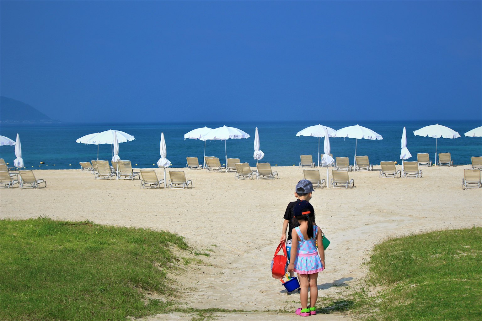 Anak-anak berjalan menuju pantai dengan payung putih dan pasir