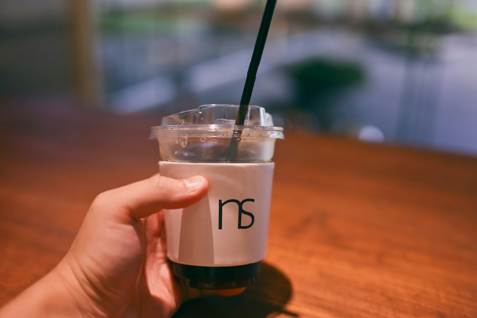 Une main tenant une tasse de café glacé avec une étiquette blanche portant un logo sur une table en bois