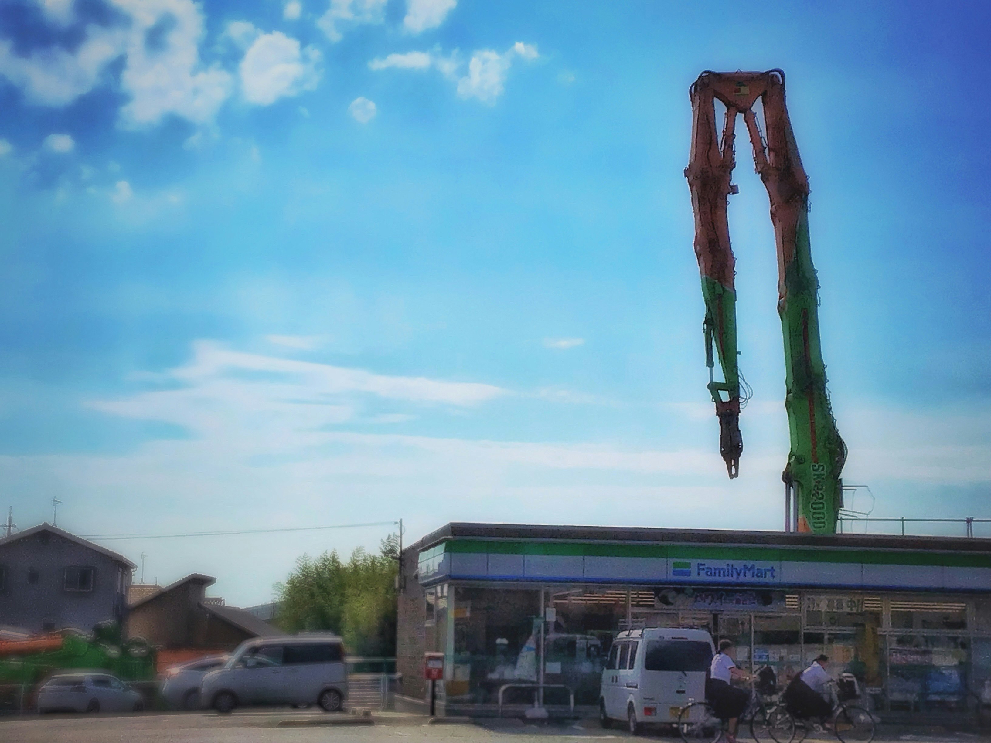 青いコンビニの裏手で伸びる巨大アームの重機のある風景