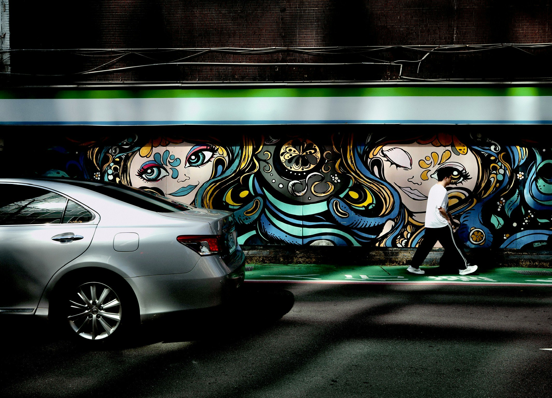 Un coche pasando junto a un mural vibrante con rostros artísticos y patrones en espiral con un peatón caminando