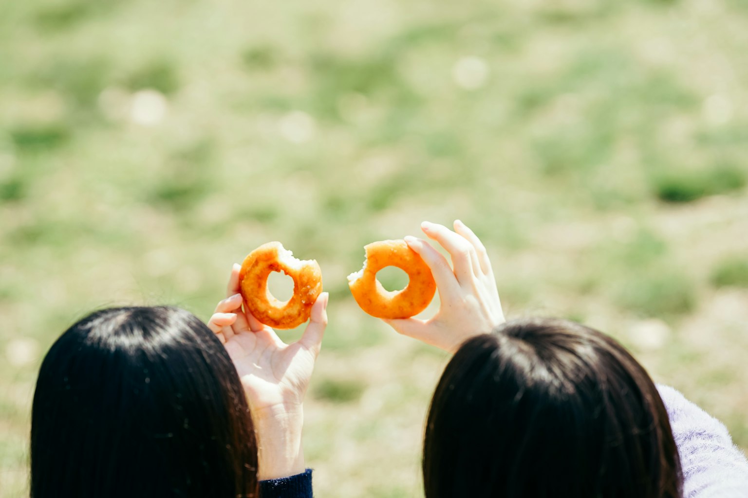 Zwei Frauen halten orangefarbene Donuts vor einem grünen Gras-Hintergrund