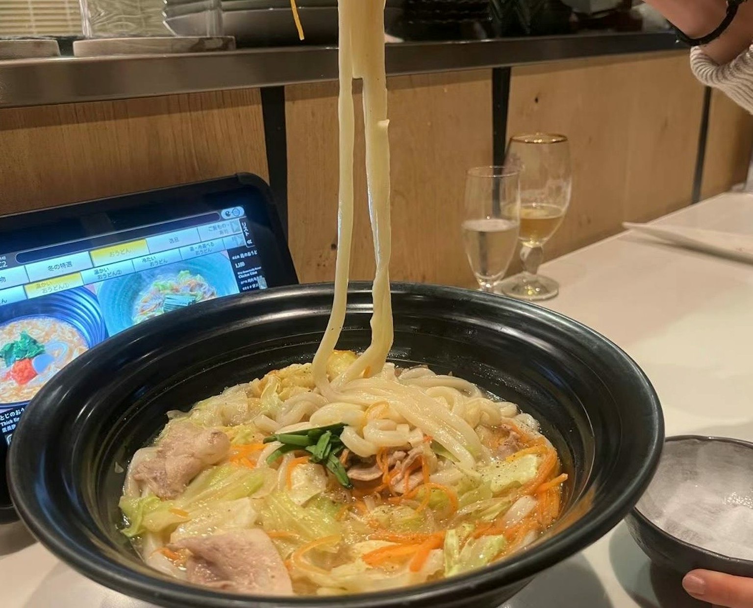 Hand lifting udon noodles from a bowl with vegetables and broth