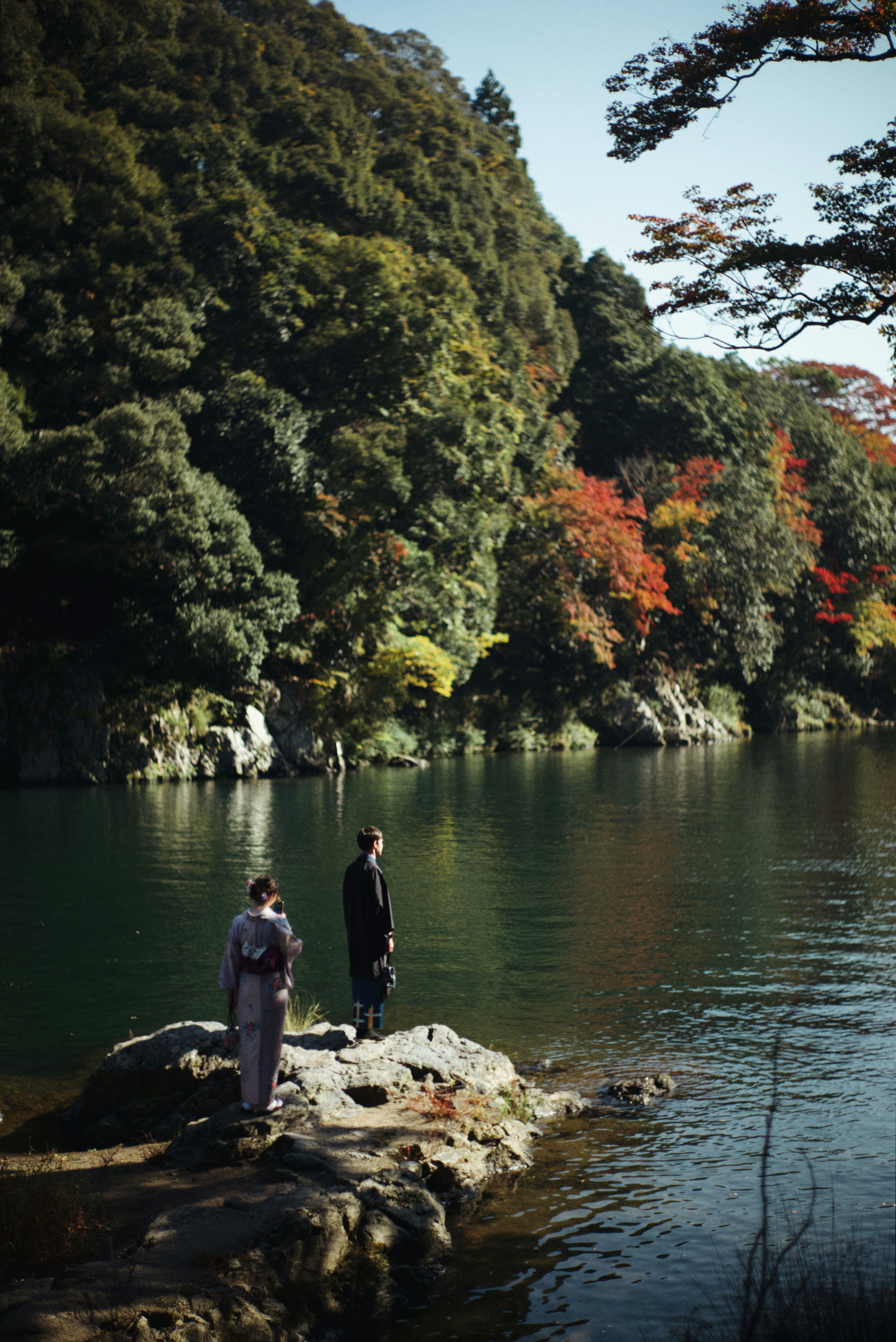 嵐山の川沿いに立つ着物姿の人と紅葉の山並み