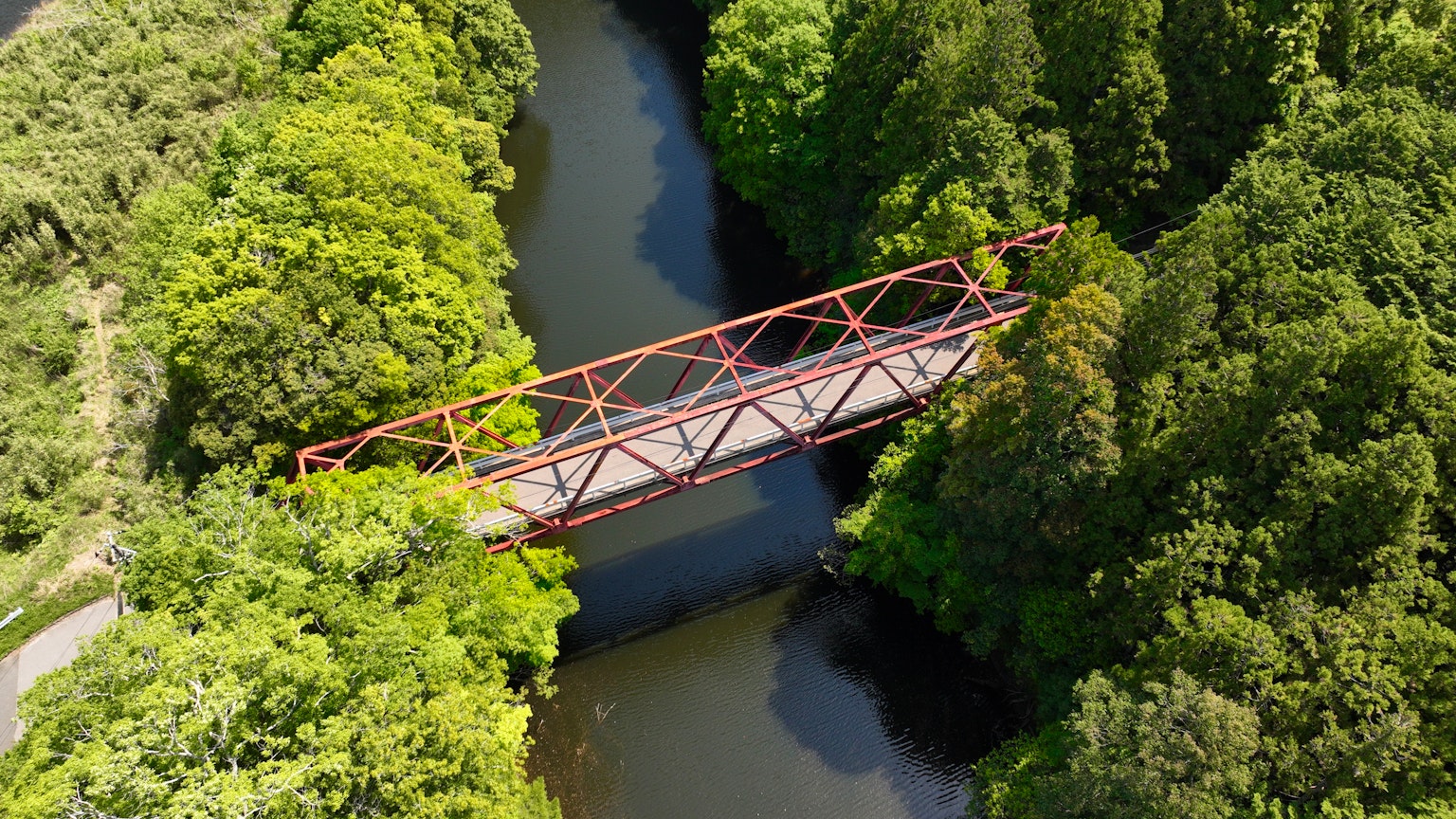 Jembatan besi merah di atas sungai dikelilingi hutan hijau yang rimbun