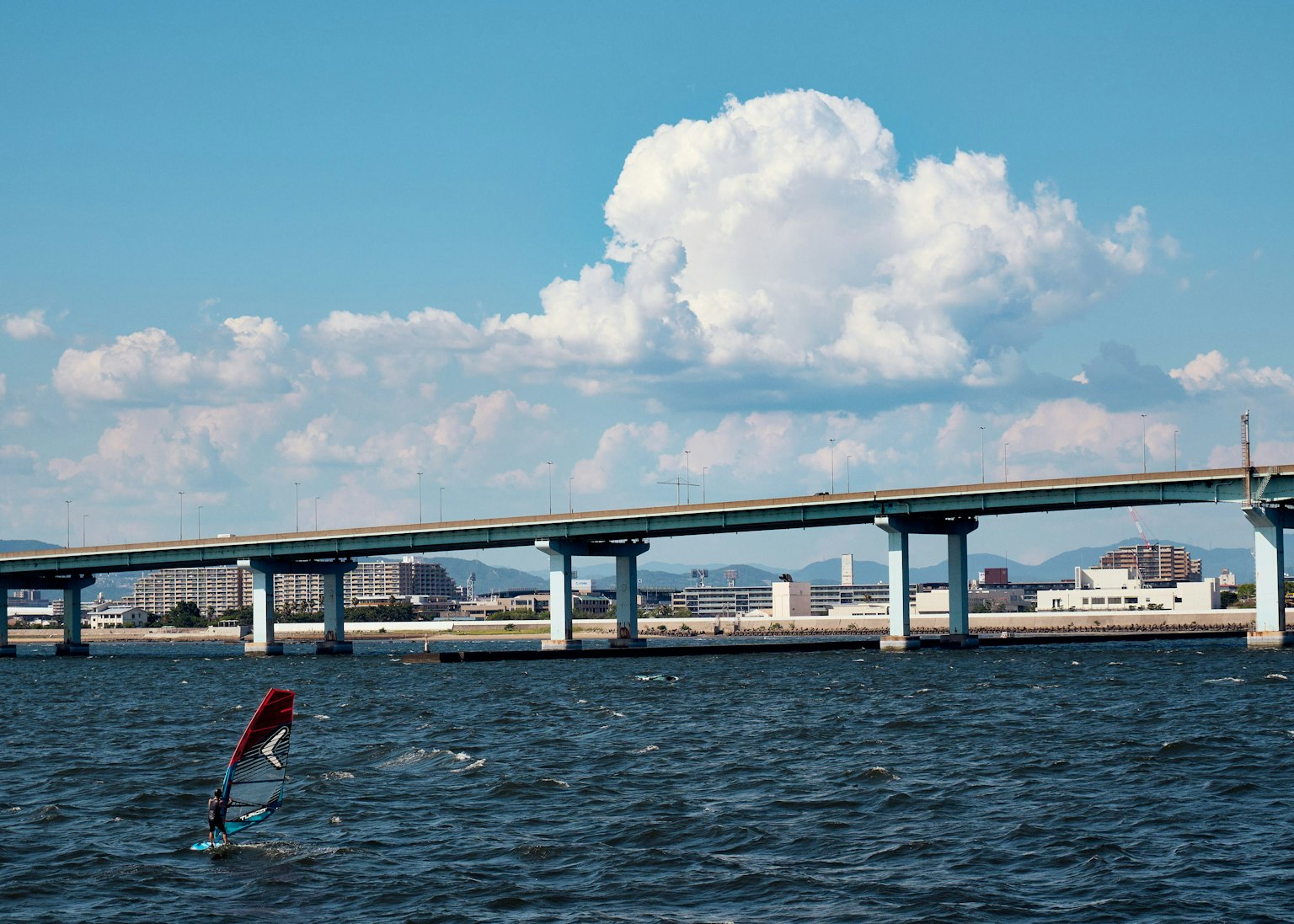Un windsurfista en una superficie de agua con un puente y un cielo azul de fondo