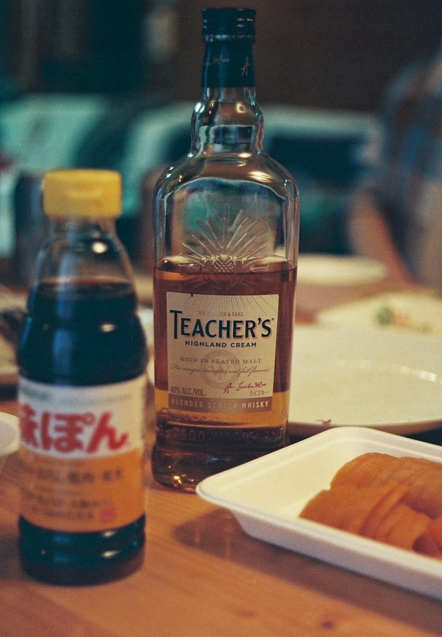 ขวดวิสกี้ที่มีป้าย Teacher's และขวดซอสถั่วเหลืองบนโต๊ะไม้พร้อมอาหาร