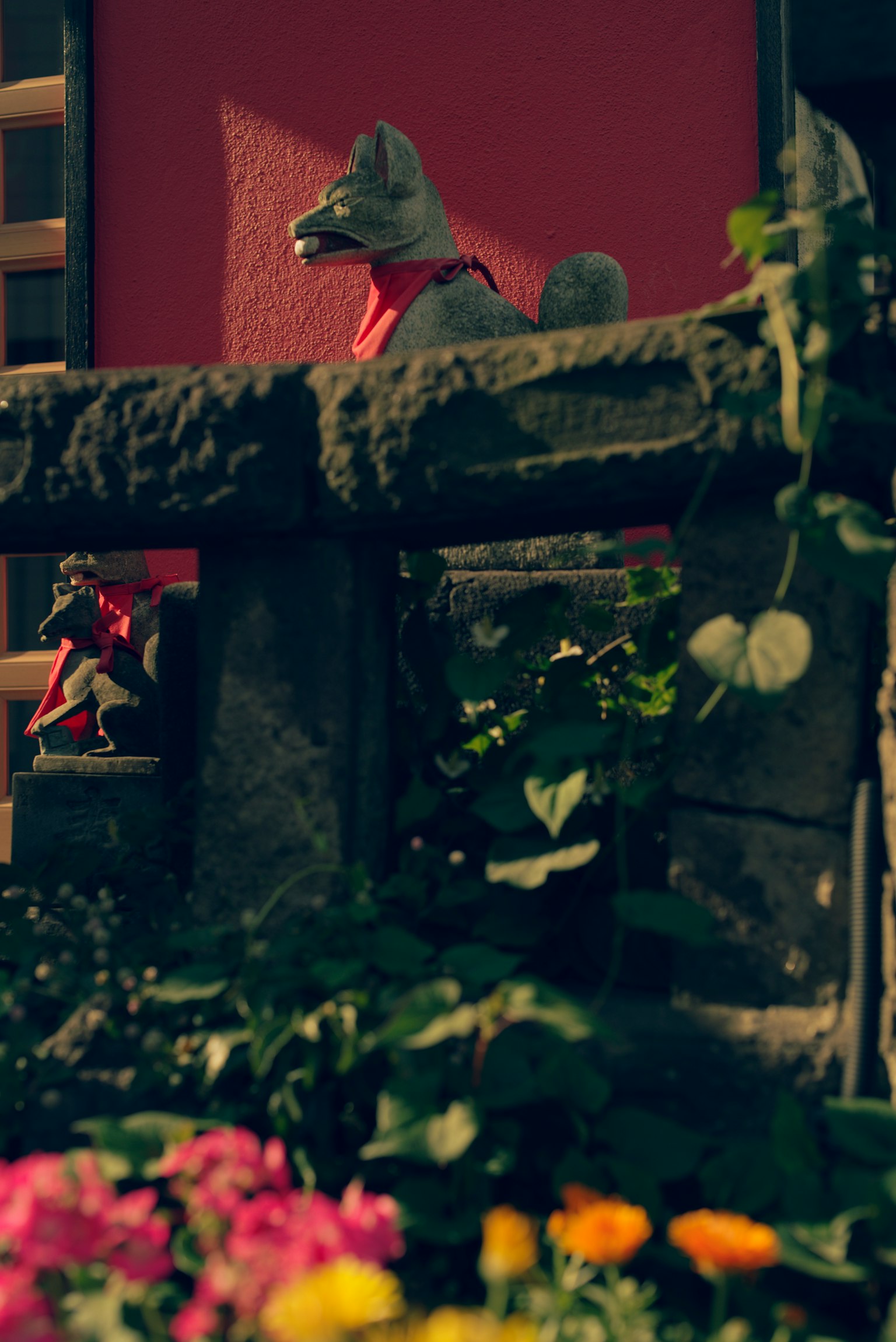 Szene mit einer grauen Hundestatue vor einer roten Wand umgeben von Blumen