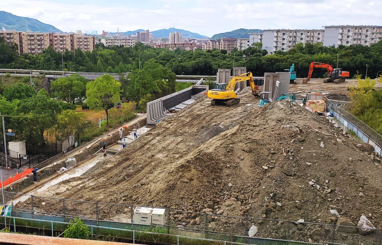 Construction site featuring heavy machinery and a dirt mound surrounded by buildings and greenery