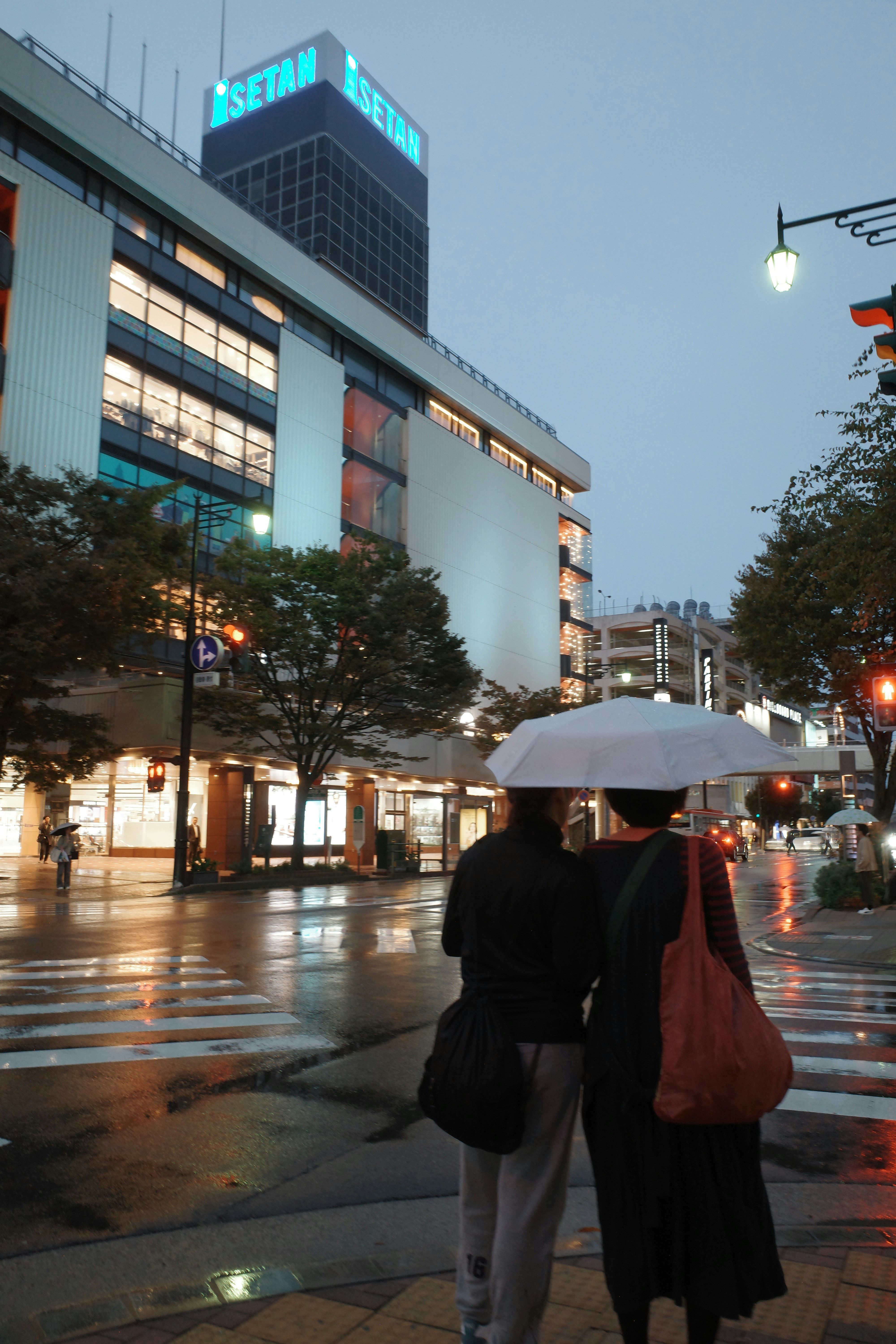 雨の夕方の駅前広場で傘をさす人々と街灯