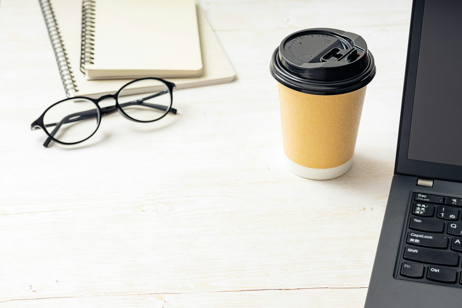 Cangkir kopi dan laptop di meja dengan buku catatan dan kacamata di dekatnya