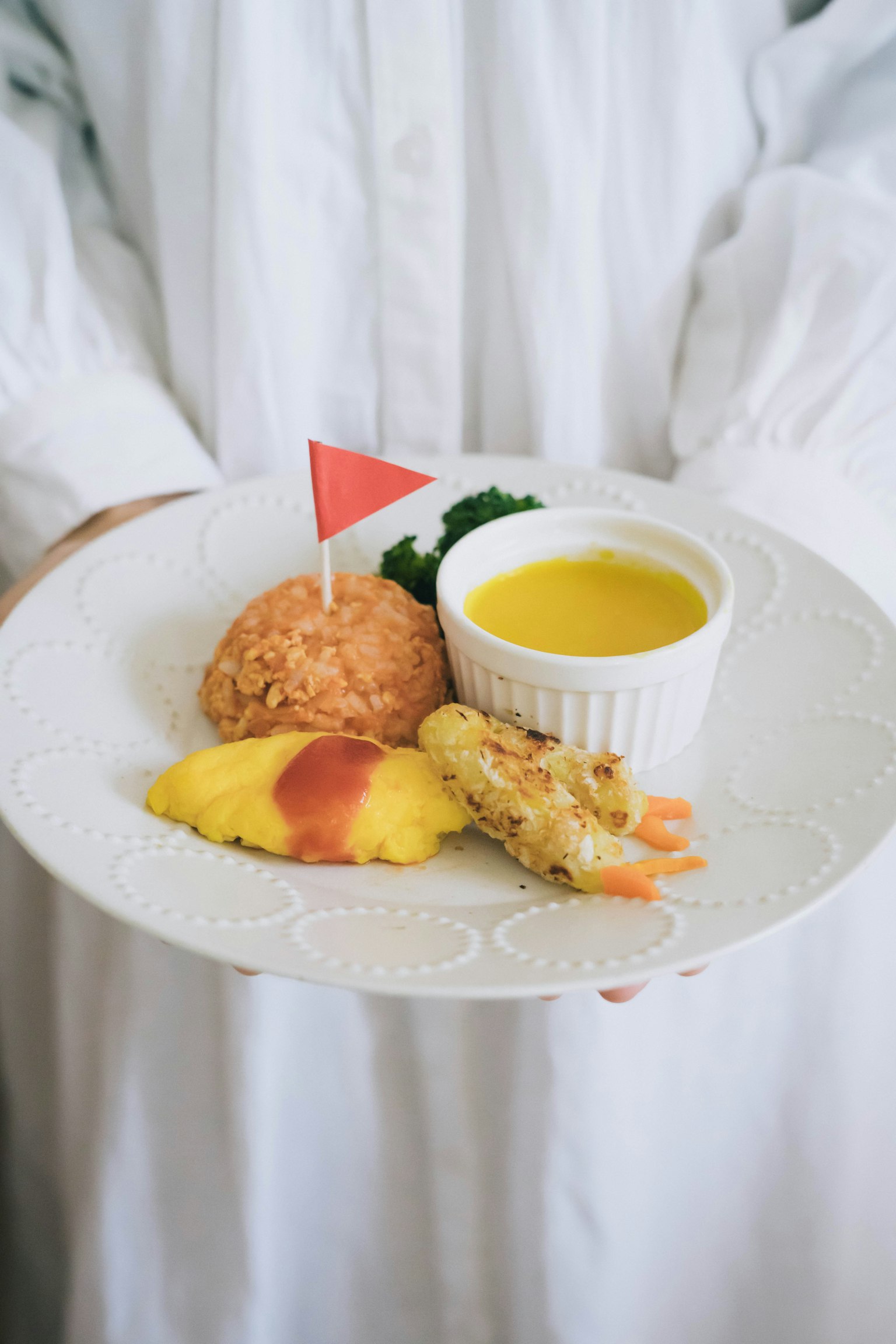 Une assiette blanche tenue à la main avec des crevettes, une omelette, du riz et un petit bol de sauce décoré d'un drapeau rouge