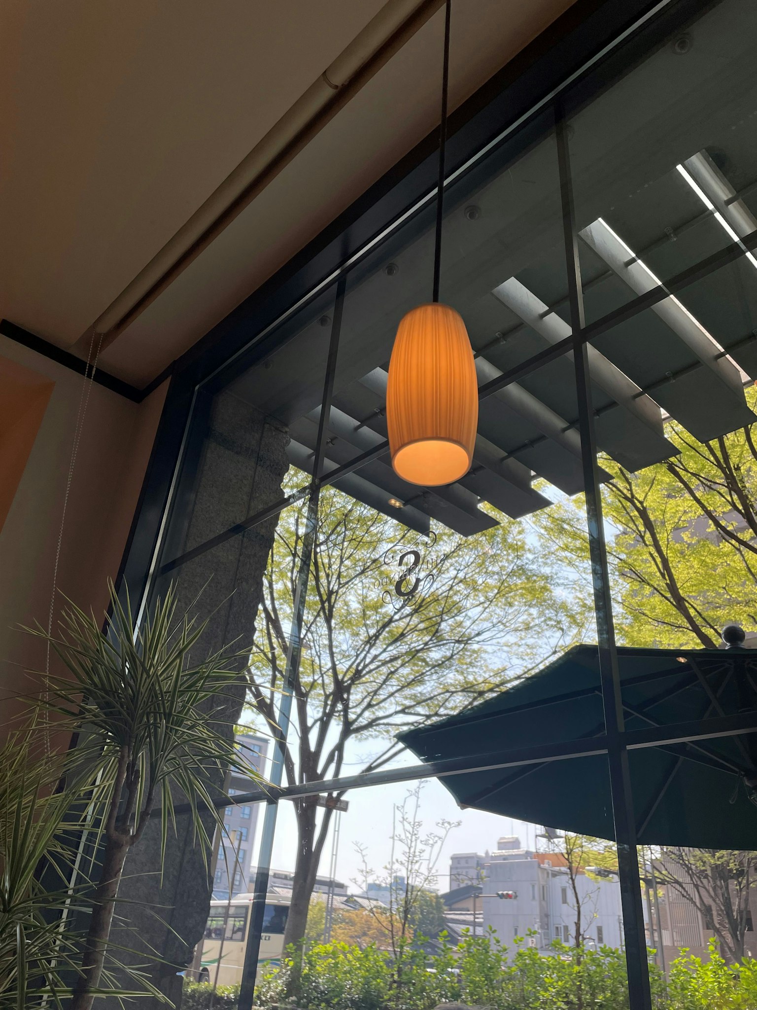 Lampu gantung oranye cerah dengan pohon hijau terlihat melalui jendela besar