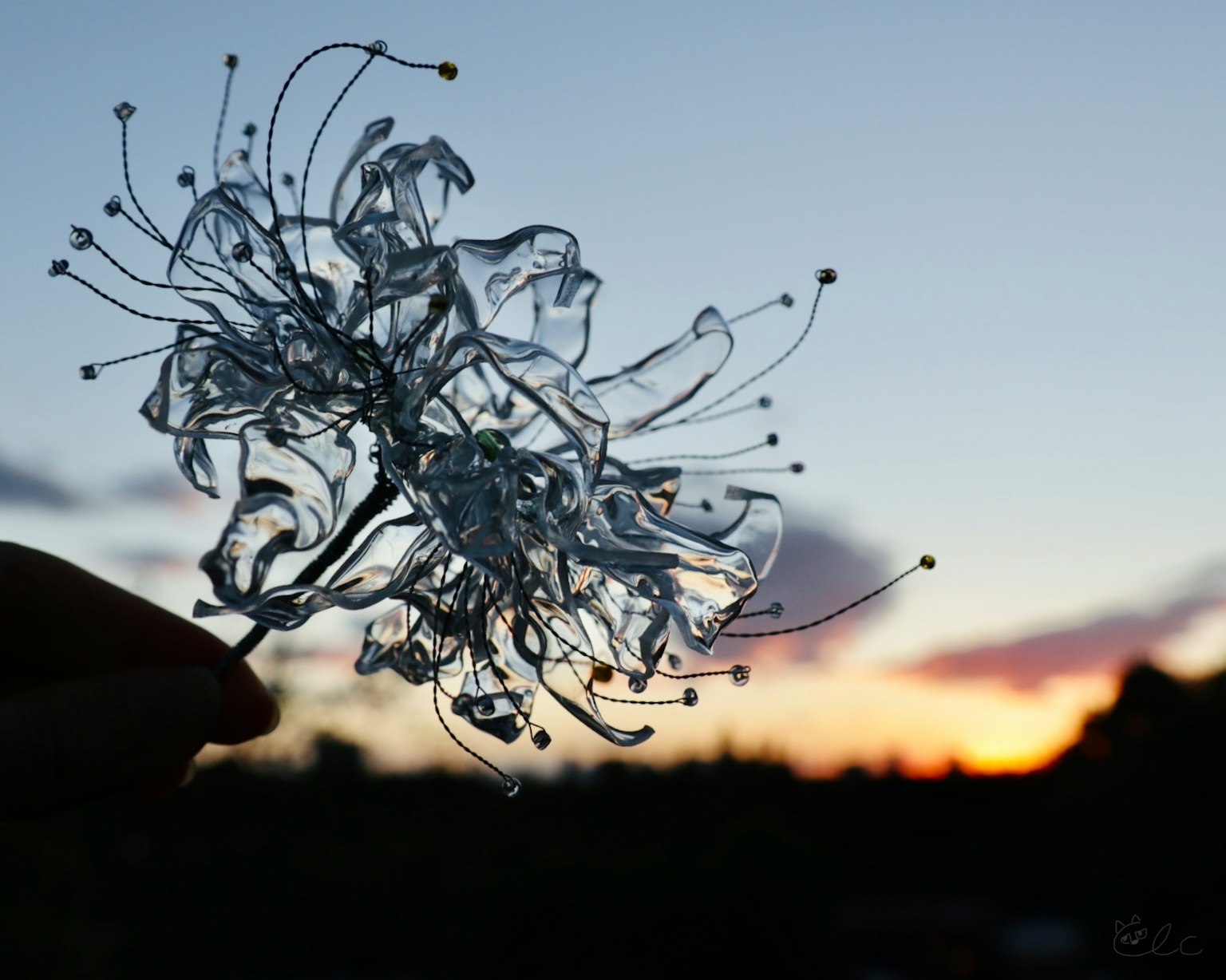 Mano sosteniendo un objeto transparente en forma de flor contra un fondo de atardecer