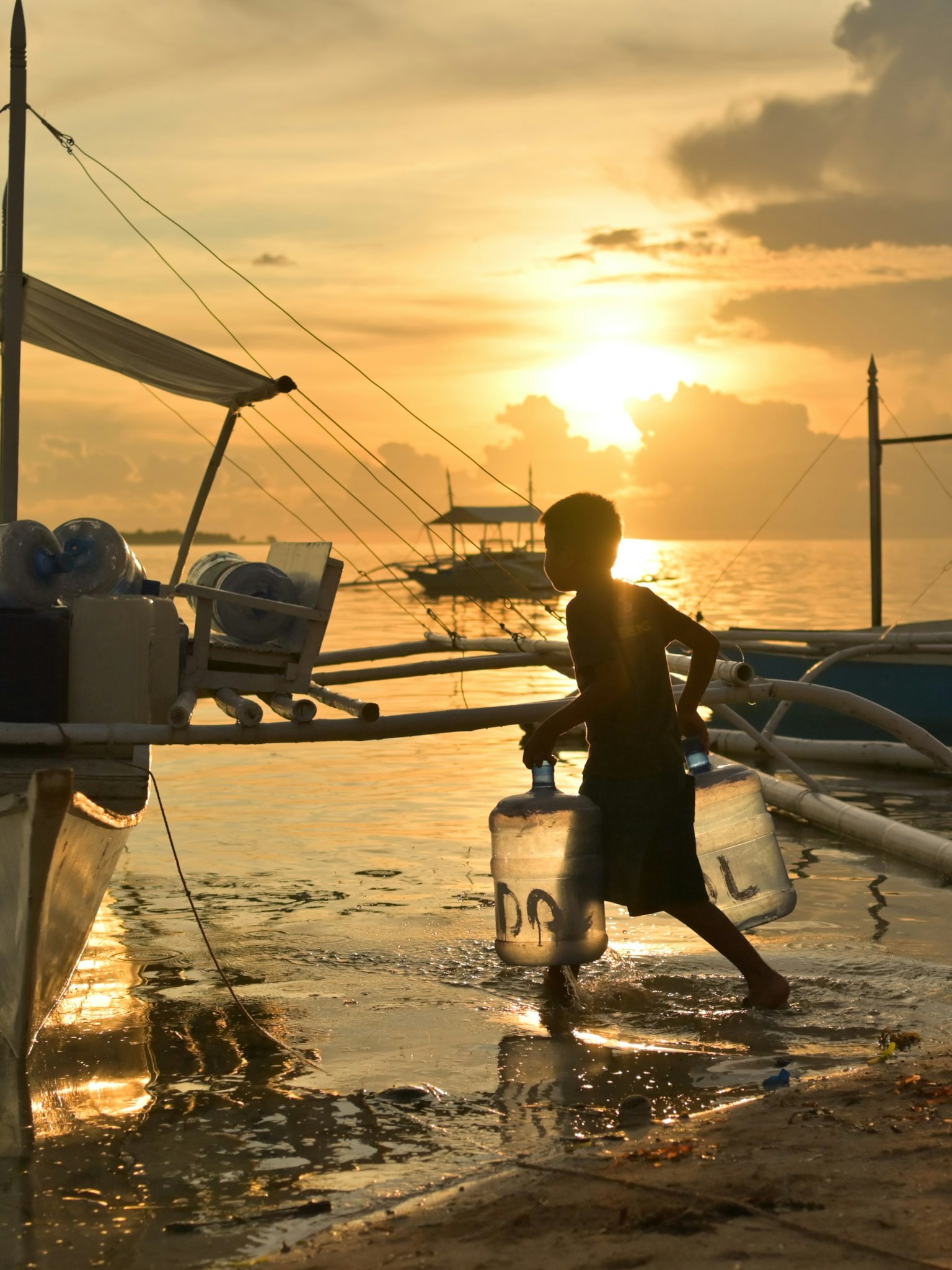 Silhouette eines Jungen, der Wasser gegen den Sonnenuntergang trägt, mit Booten und Meer im Hintergrund