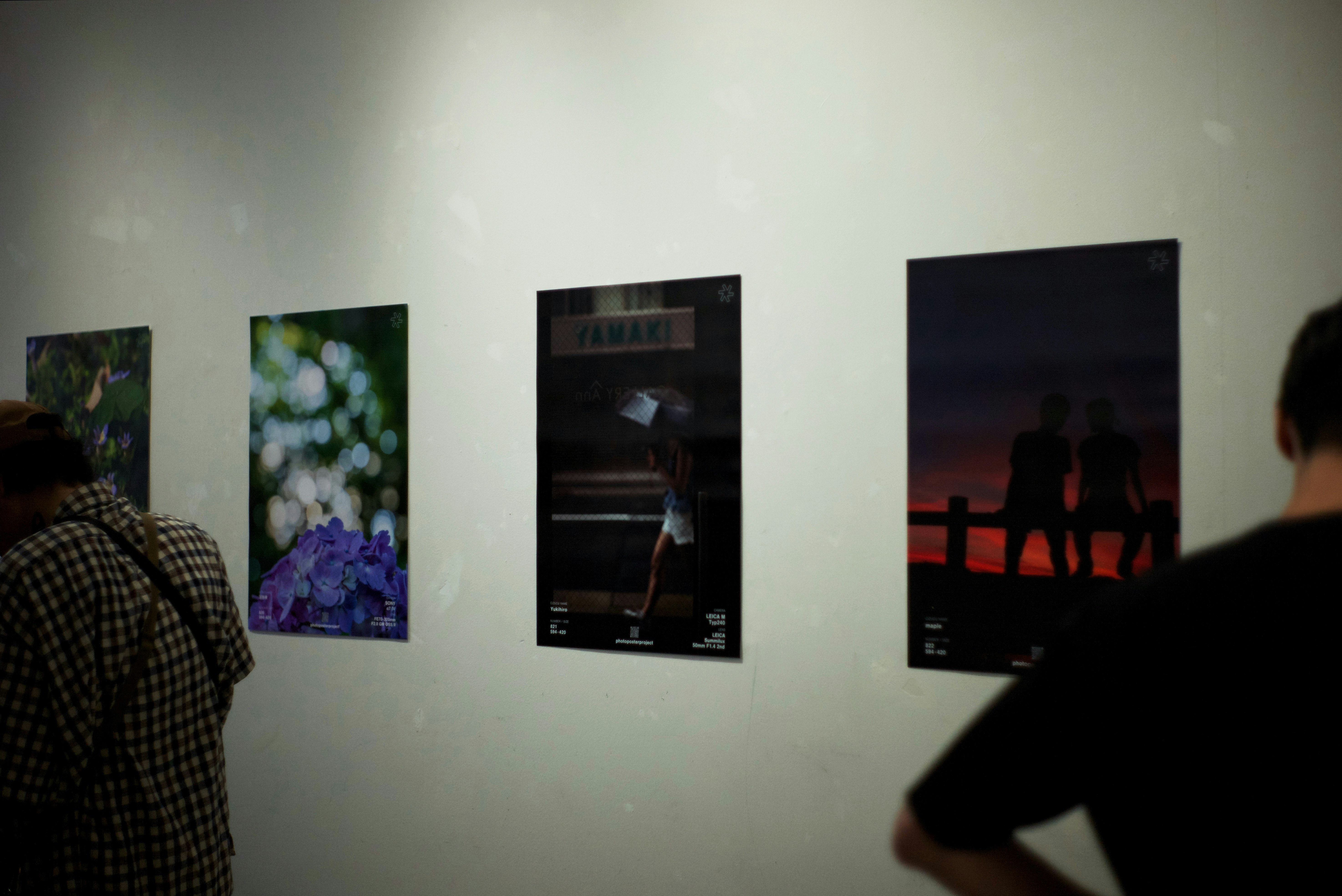 poster fotografici esposti su una parete di galleria con visitatori di spalle
