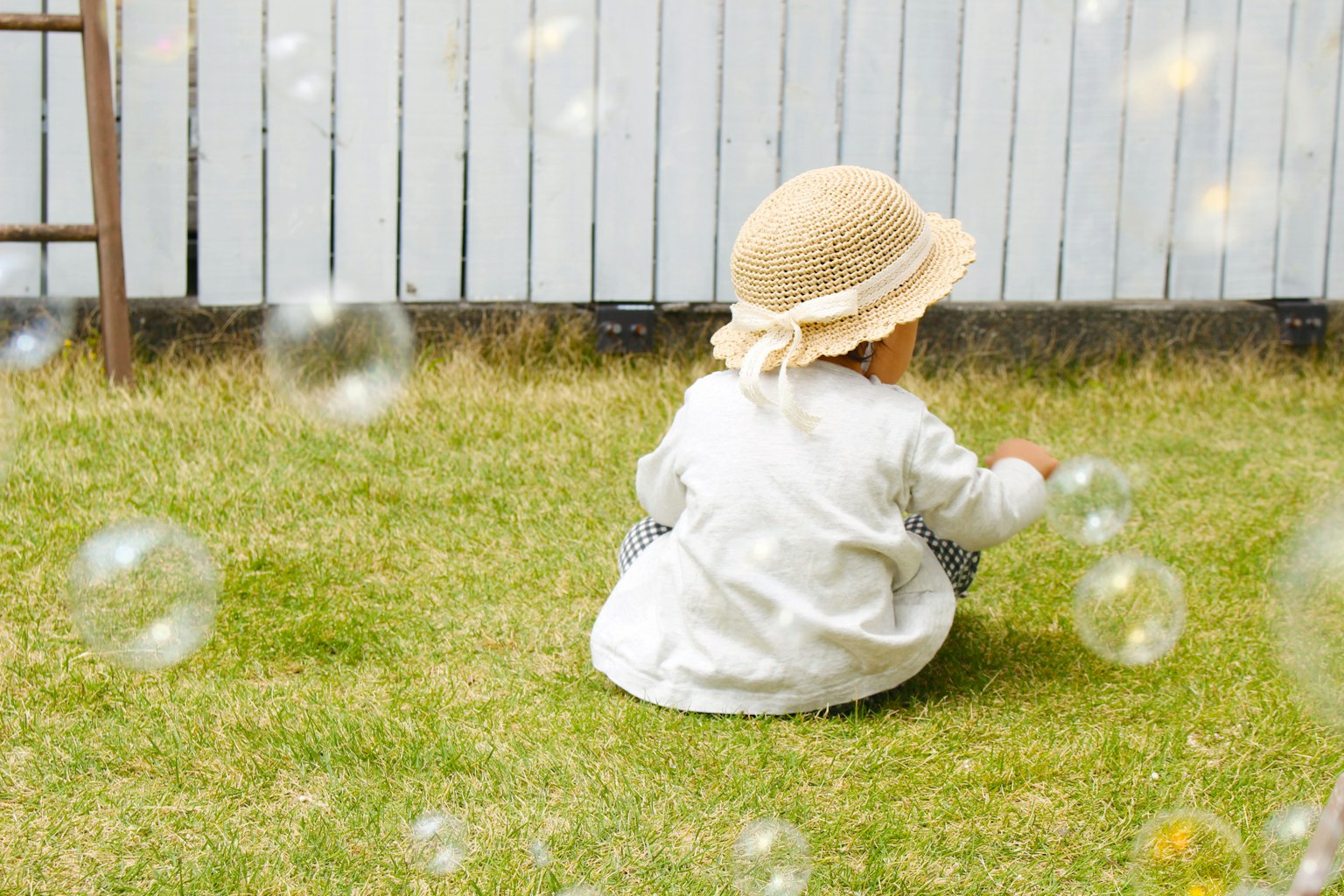 Ein Kind spielt zwischen Seifenblasen von hinten gesehen in einem weißen Hemd und einem Strohhut