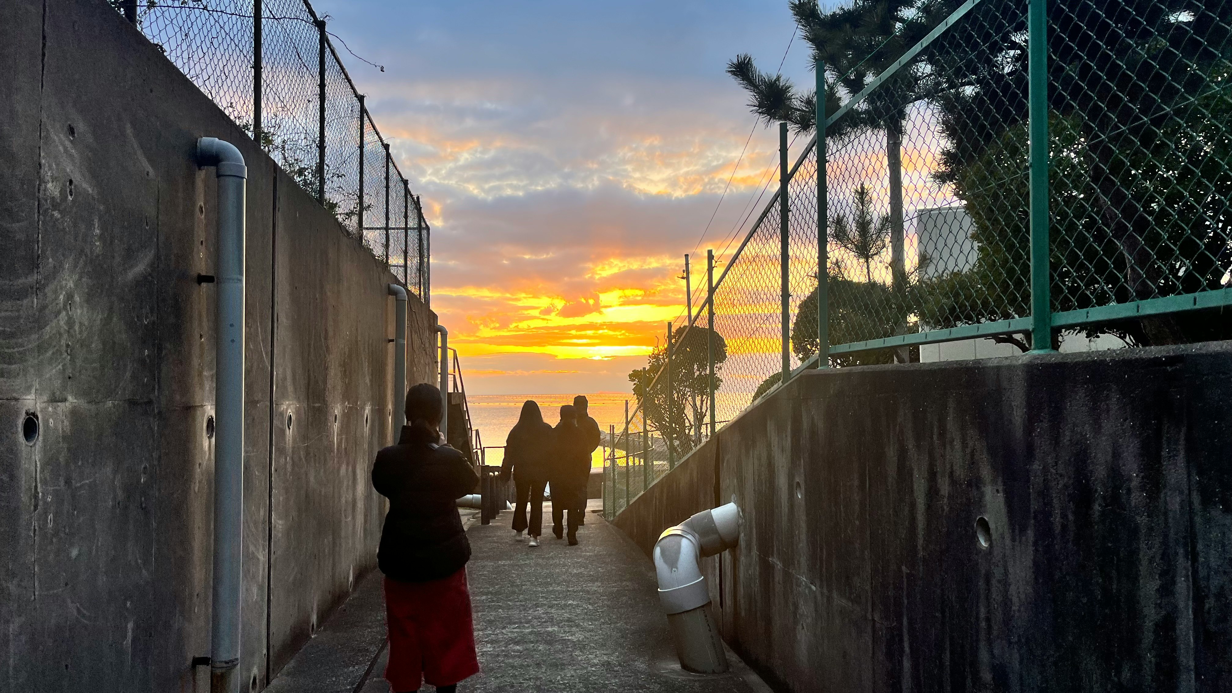 フェンスに挟まれた通路を歩く人々と夕焼けの空