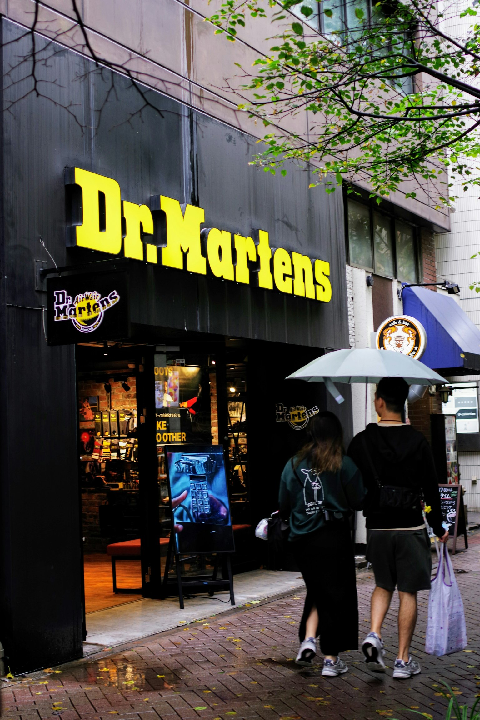 Pasangan berjalan di depan toko Dr Martens pada hari hujan sambil memegang payung