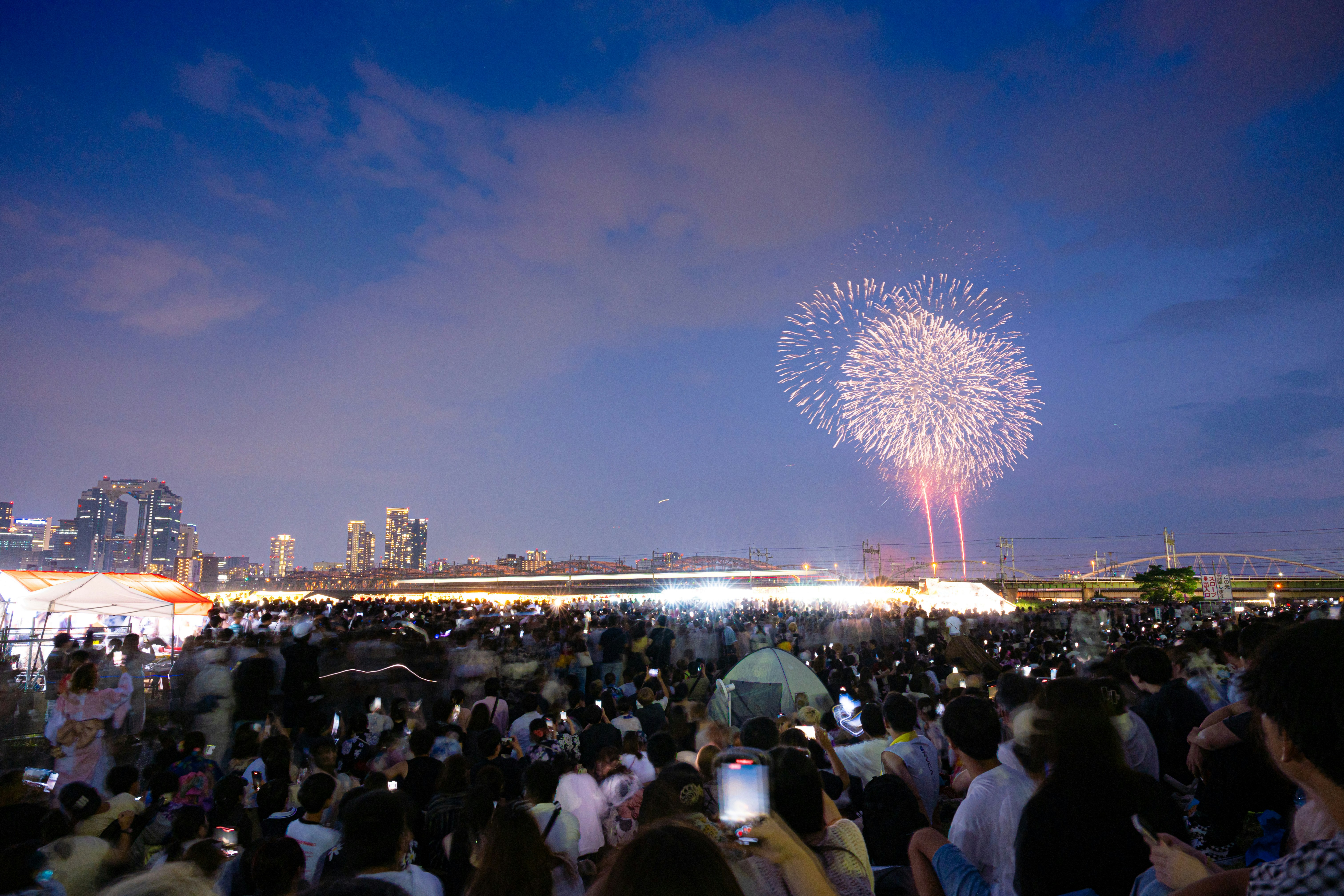 花火を見上げる人々が集まる河川敷の大規模イベント会場