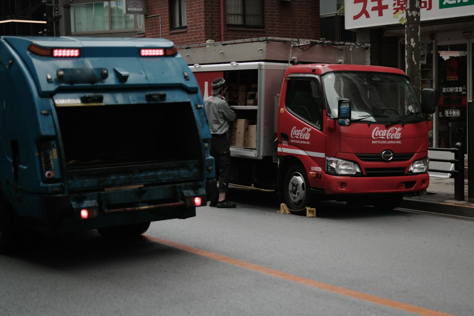 파란 쓰레기 트럭과 빨간 코카콜라 배달 트럭이 도시 거리에서 주차되어 있다