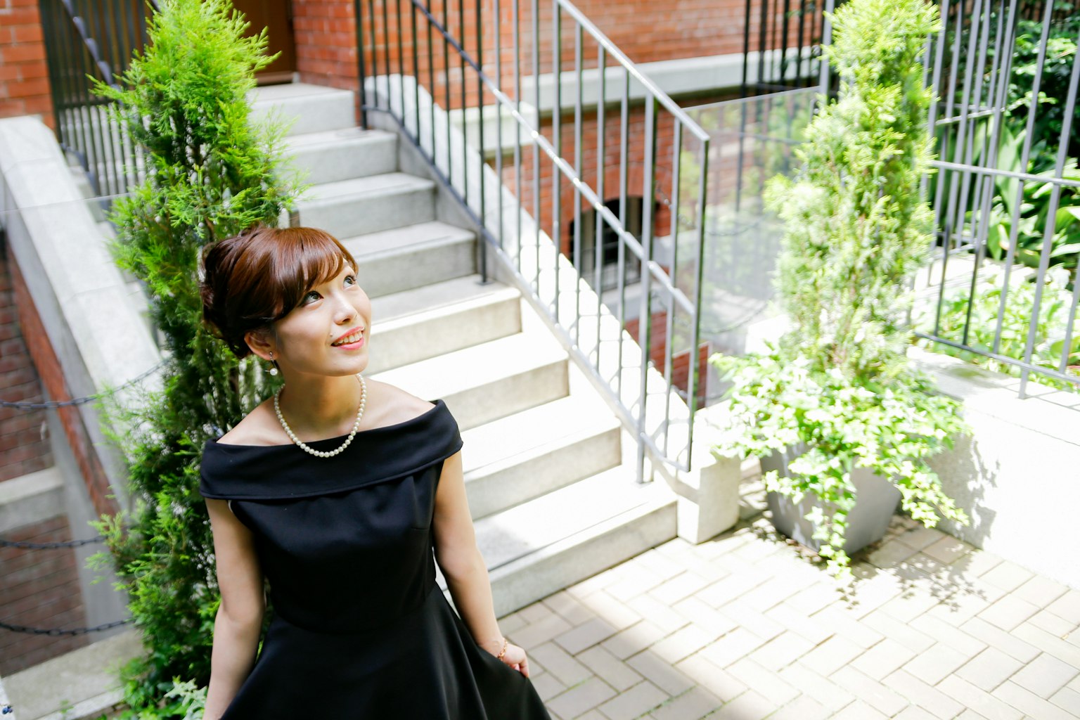 Femme en robe noire debout près d'un escalier souriante entourée de plantes vertes