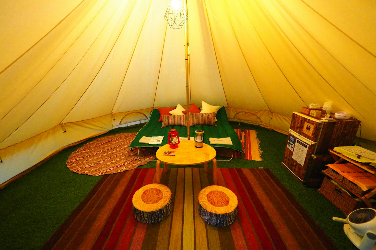 Interno accogliente di una tenda con tappeti colorati, un tavolo in legno con sgabelli e letti con cuscini morbidi