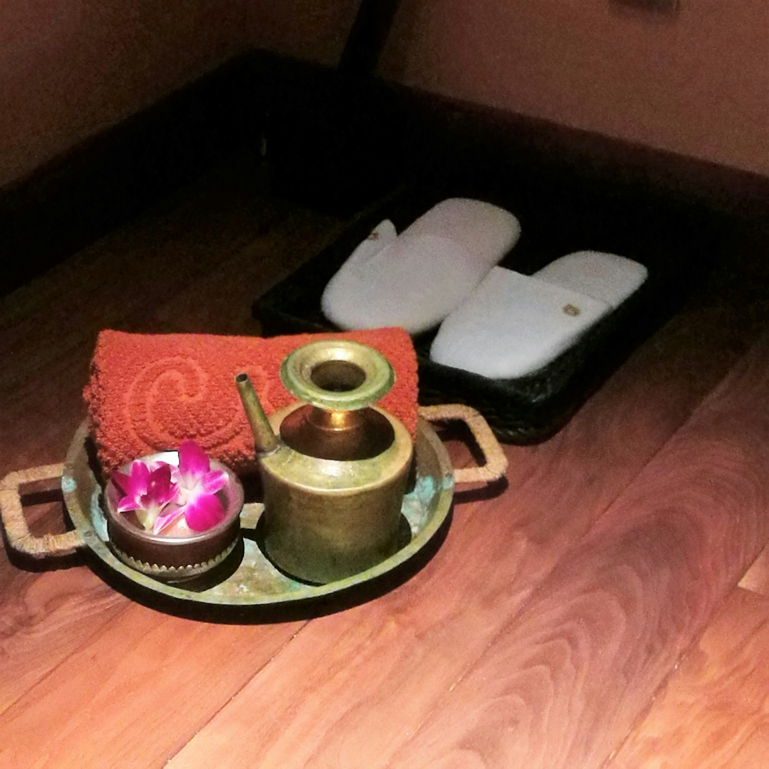 Un sol en bois présente un plateau avec une théière en métal et un bol avec une fleur, accompagné d'un tissu rouge et de chaussons