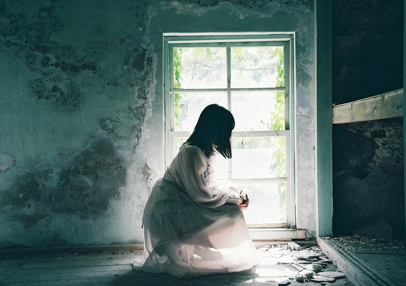 Una donna seduta vicino a una finestra in una stanza poco illuminata che indossa un vestito bianco luce che entra dalla finestra con ombre verdi