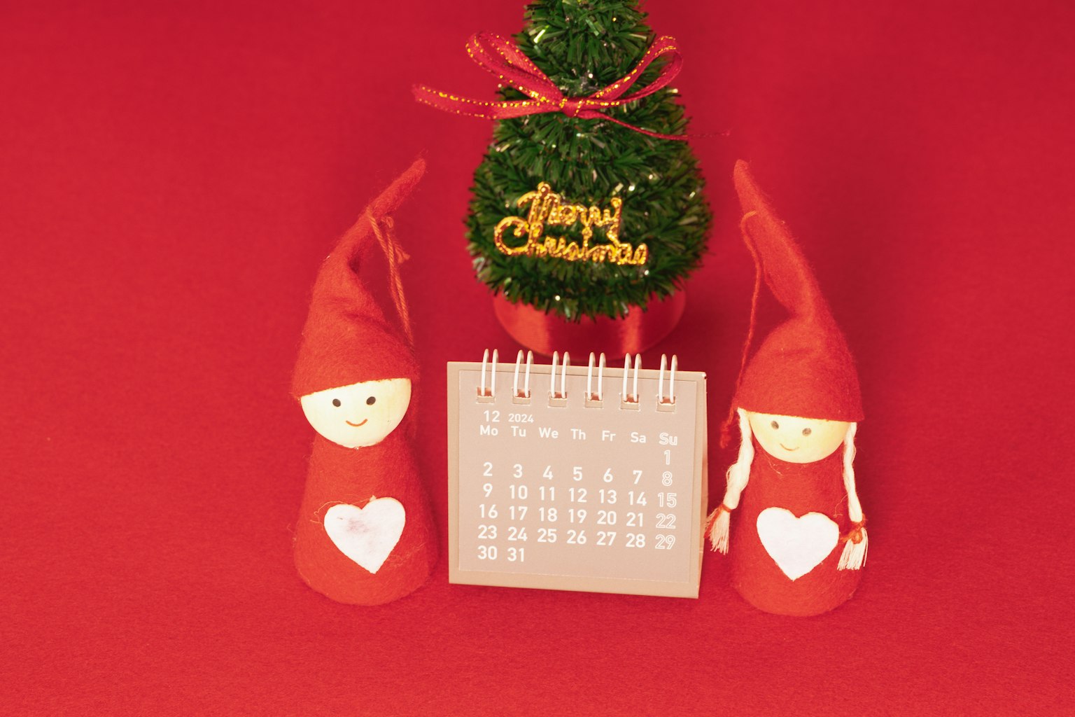 Zwei rotmützige Puppen mit Herzen neben einem Kalender und einem kleinen Weihnachtsbaum auf rotem Hintergrund