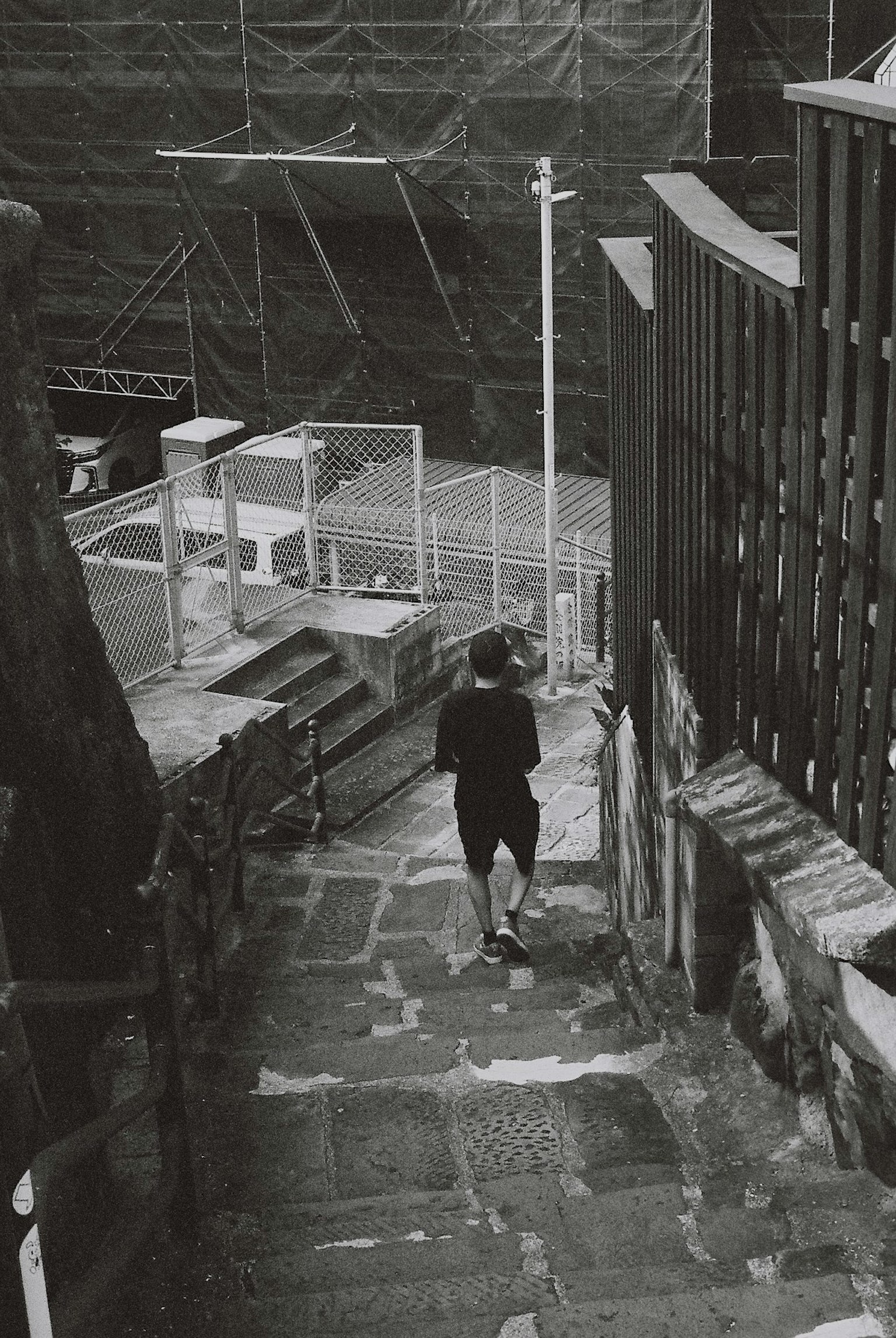 Black and white photo of a person walking down stairs with a fence and construction site in the background
