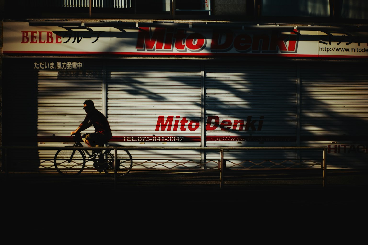 自転車に乗る人物が影の中を通り過ぎる Mito Denki のシャッターが閉まった店舗