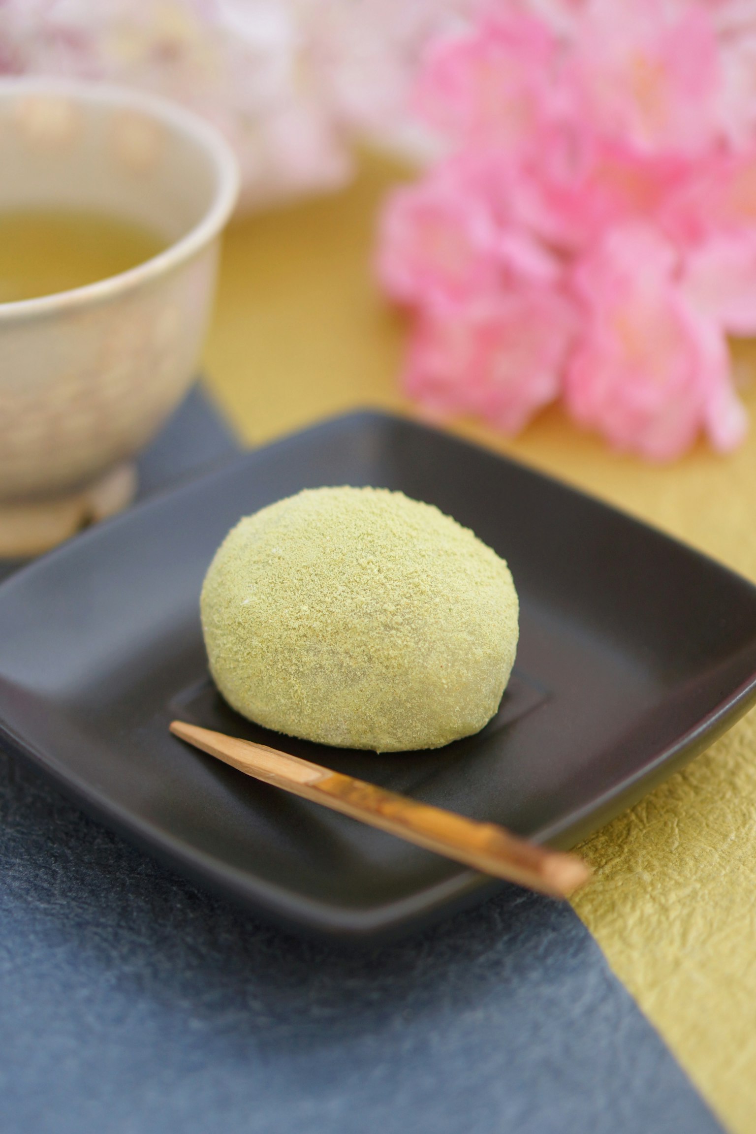 Mochi al matcha su un piatto nero con un bastoncino di legno accanto a una tazza di tè verde e fiori di ciliegio sullo sfondo