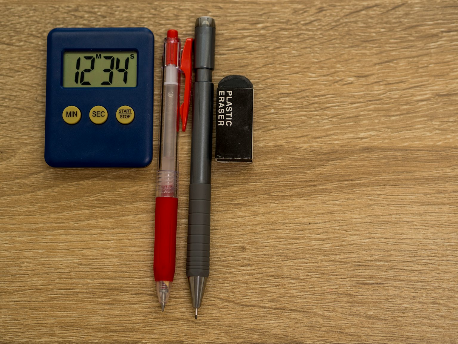 Ein blauer digitaler Timer neben einem roten Stift, einem grauen Stift und einem schwarzen Radiergummi auf einem Holztisch
