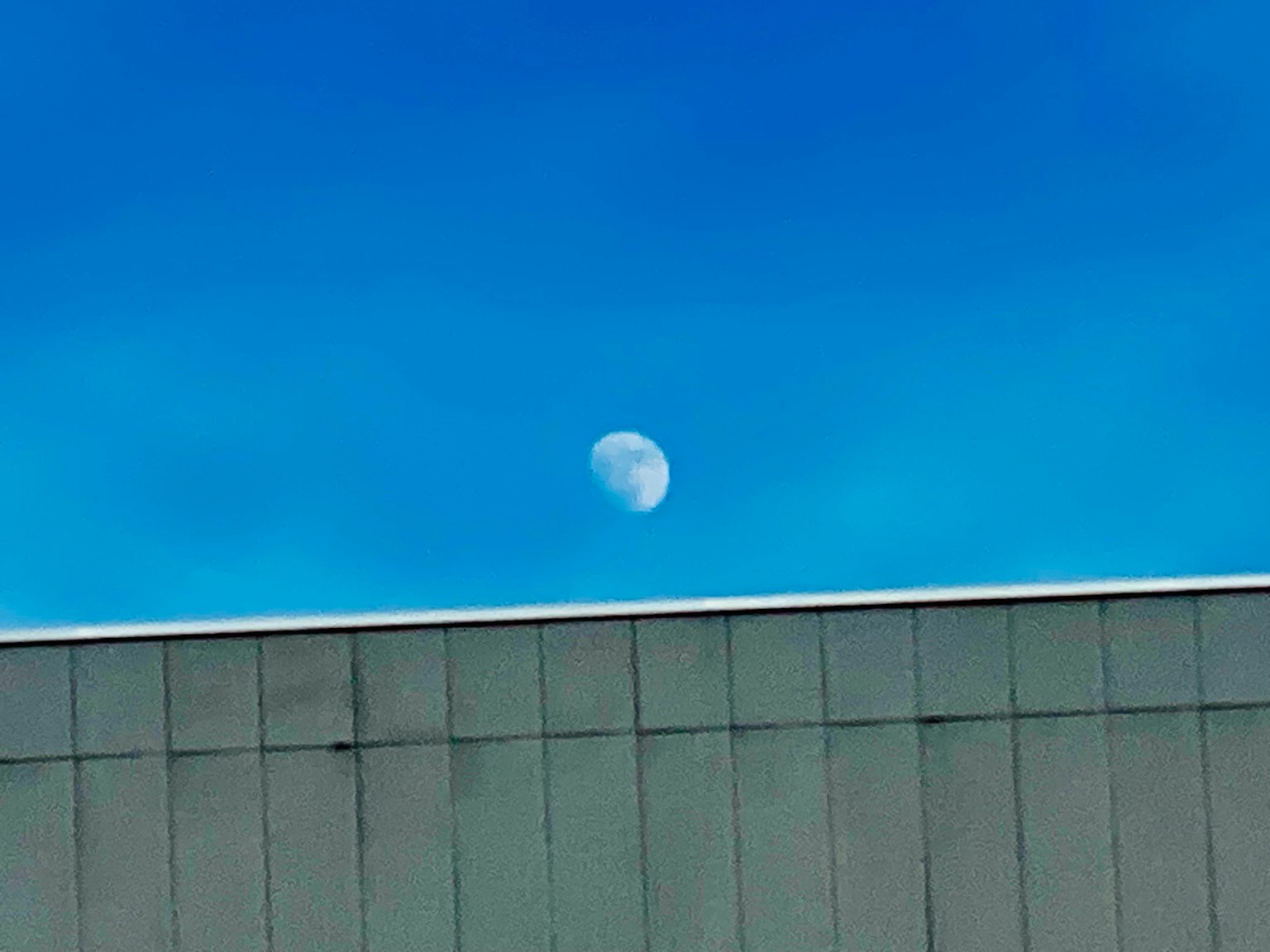 Luna visibile contro un cielo blu chiaro sopra un tetto di edificio