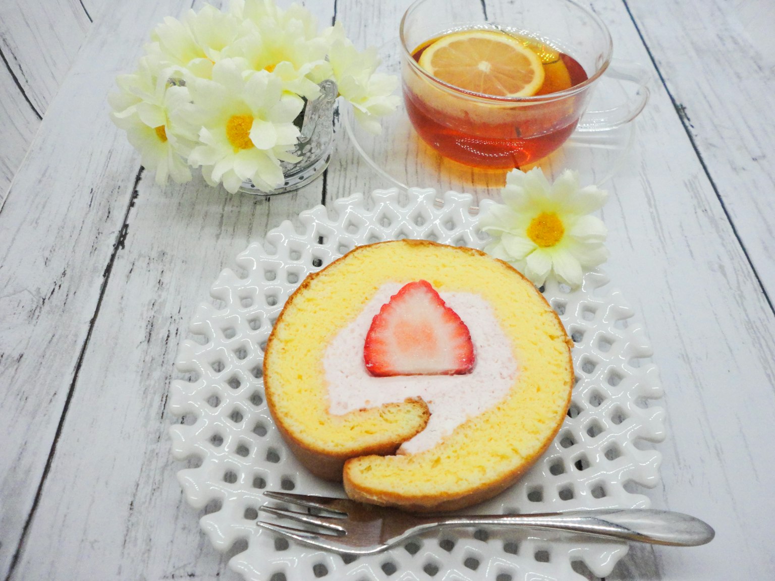 Gâteau roulé jaune avec fraise sur le dessus disposé à côté de fleurs blanches et de thé