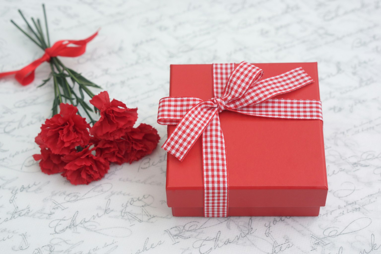 Hộp quà màu đỏ với ruy băng kẻ ô bên cạnh bó hoa cẩm chướng đỏ