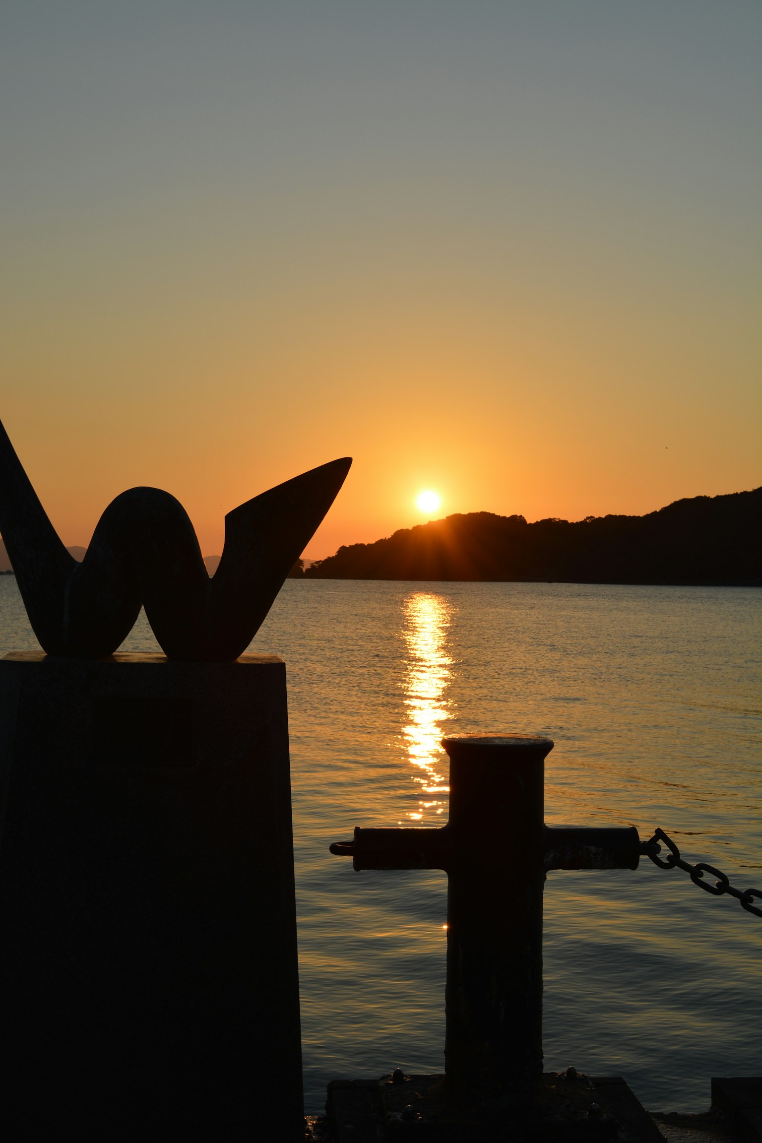 Silueta de una cruz a la derecha y una escultura 'W' a la izquierda con la puesta de sol sobre el mar