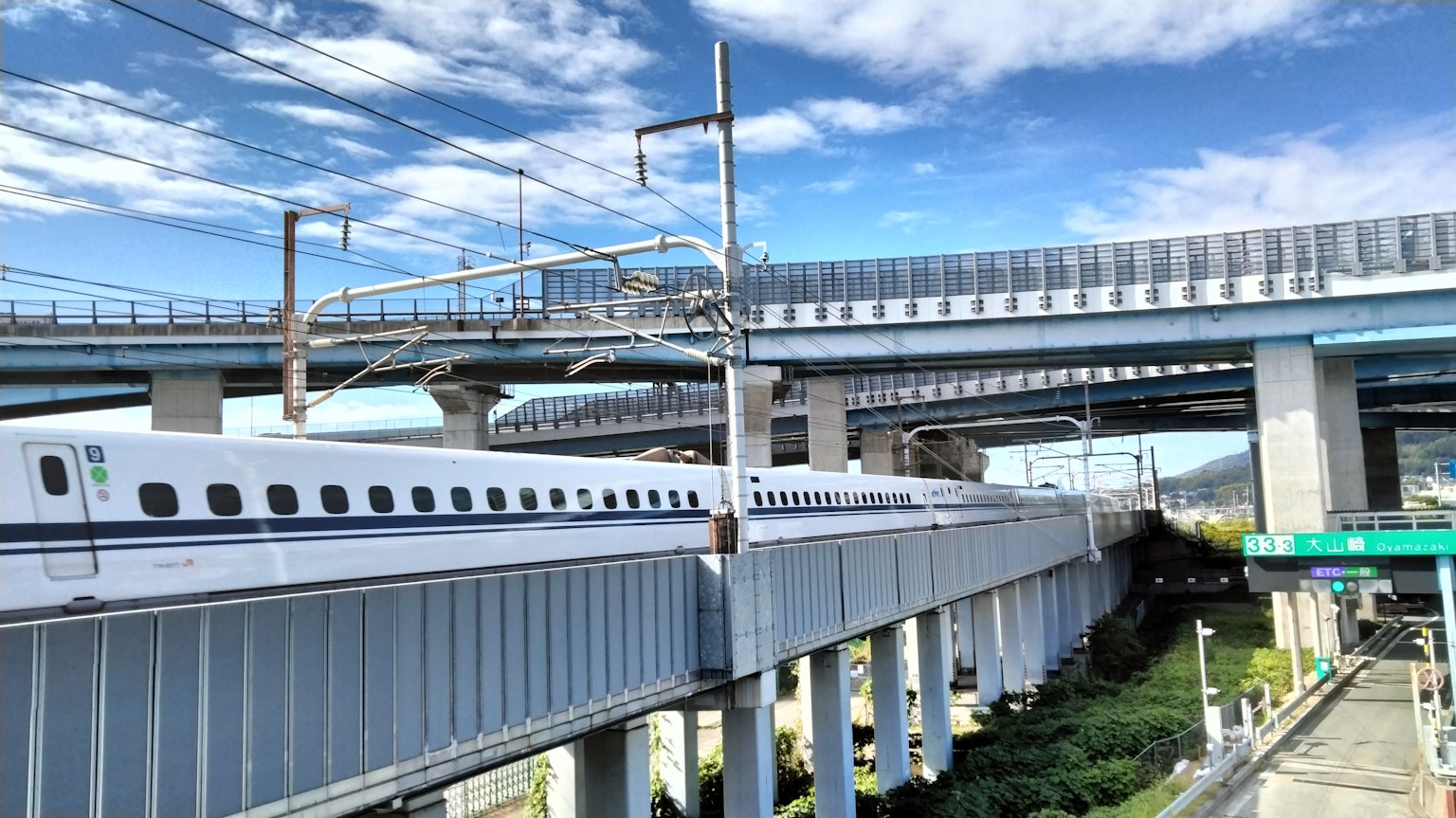 Shinkansen đi qua dưới cầu vượt với bầu trời xanh và mây ở phía sau
