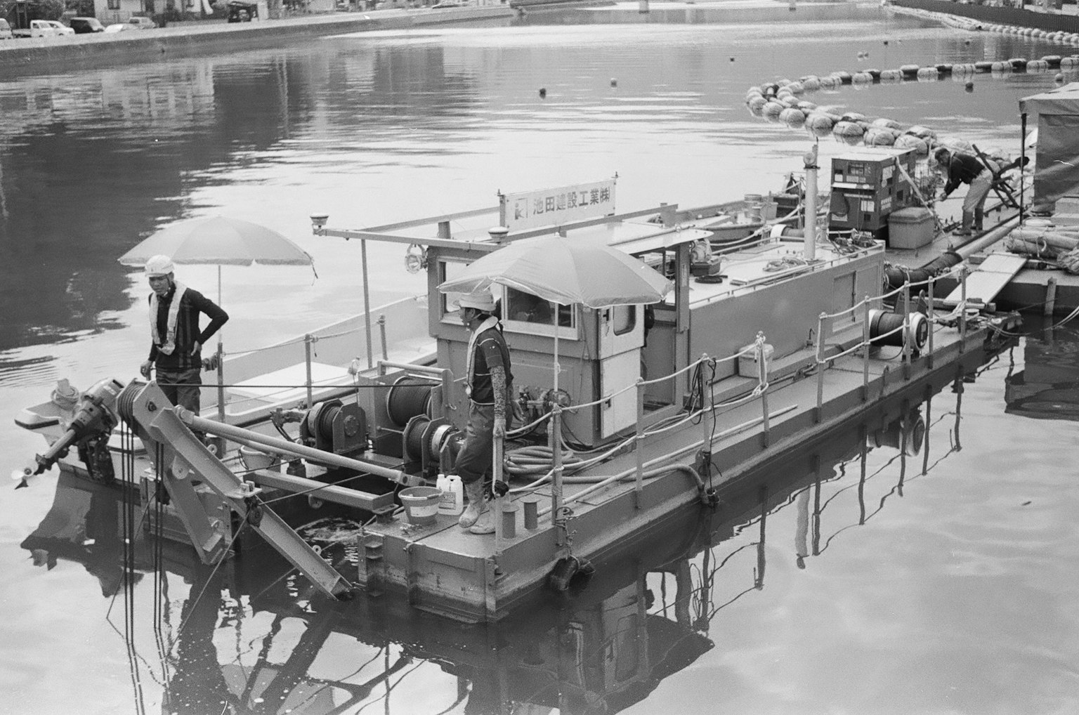 ภาพขาวดำของเรือทำงานจอดอยู่บนผิวน้ำที่สงบพร้อมคนงานยืนอยู่ข้างๆ