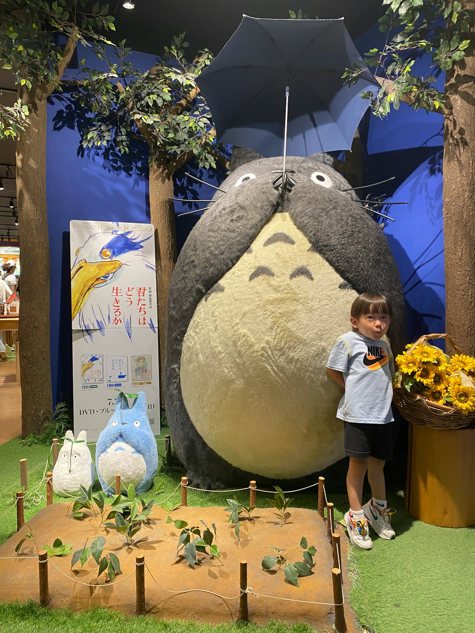 A child standing next to a large Totoro plush surrounded by greenery and trees